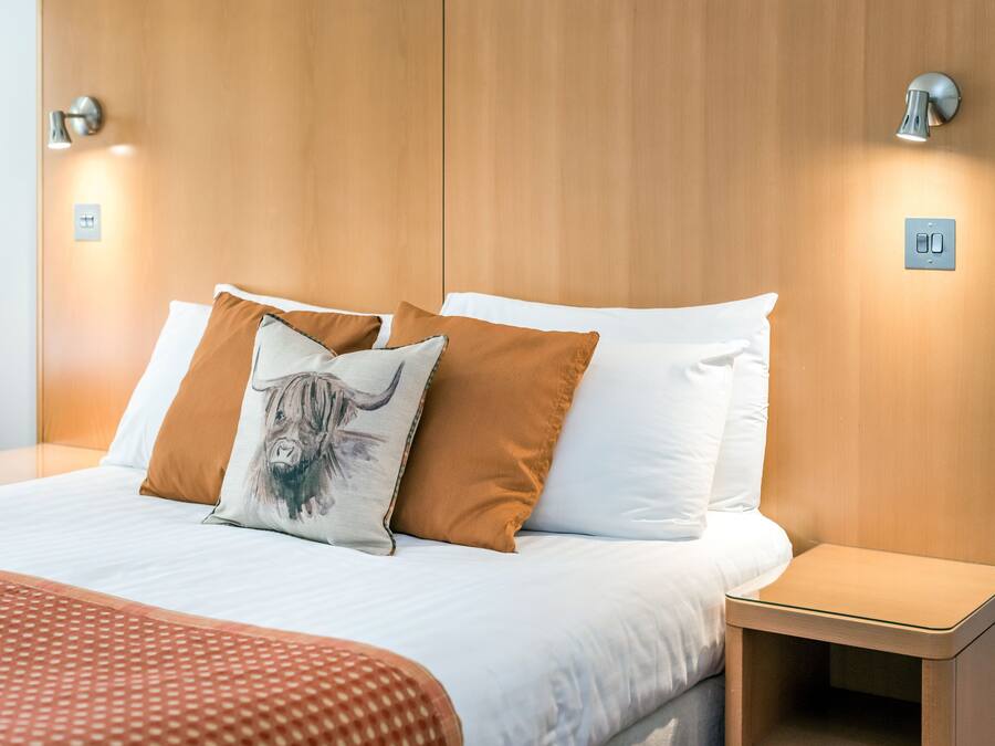 A neatly made bed with white and brown bedding, two wall-mounted light fixtures, and a small bedside table.