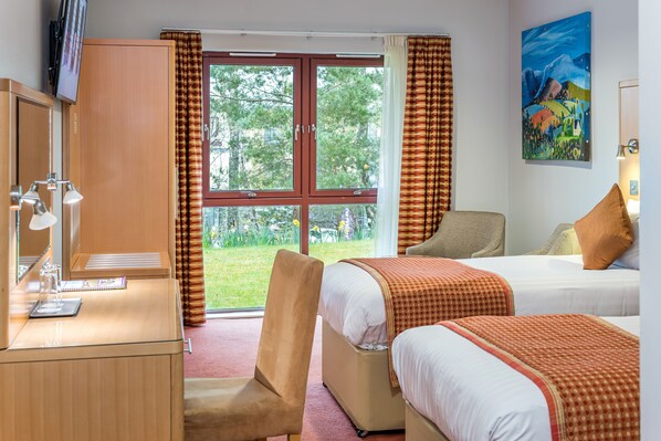 Habitación de hotel con escritorio, dos camas, una silla y un ventanal grande con cortinas.