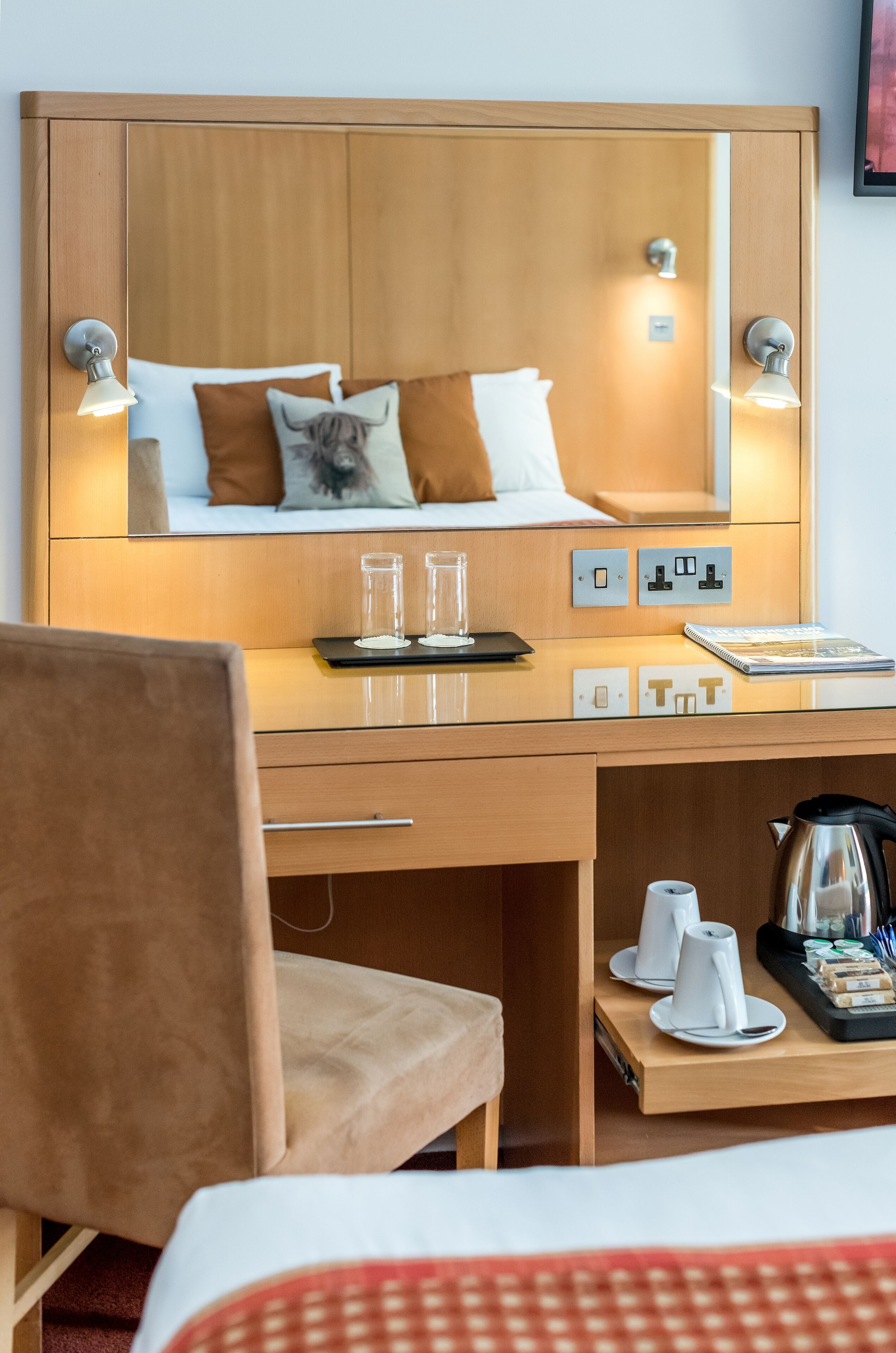 Hotelkamer met een houten hoofdbord, een bed met bruine en witte kussens, een bureau met een laptop, brillen en een notitieboekje. Er is een stoel, een koffiezetapparaat en een dienblad met kopjes en snacks.