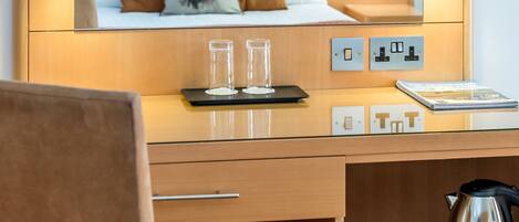Hotelkamer met een houten hoofdbord, een bed met bruine en witte kussens, een bureau met een laptop, brillen en een notitieboekje. Er is een stoel, een koffiezetapparaat en een dienblad met kopjes en snacks.
