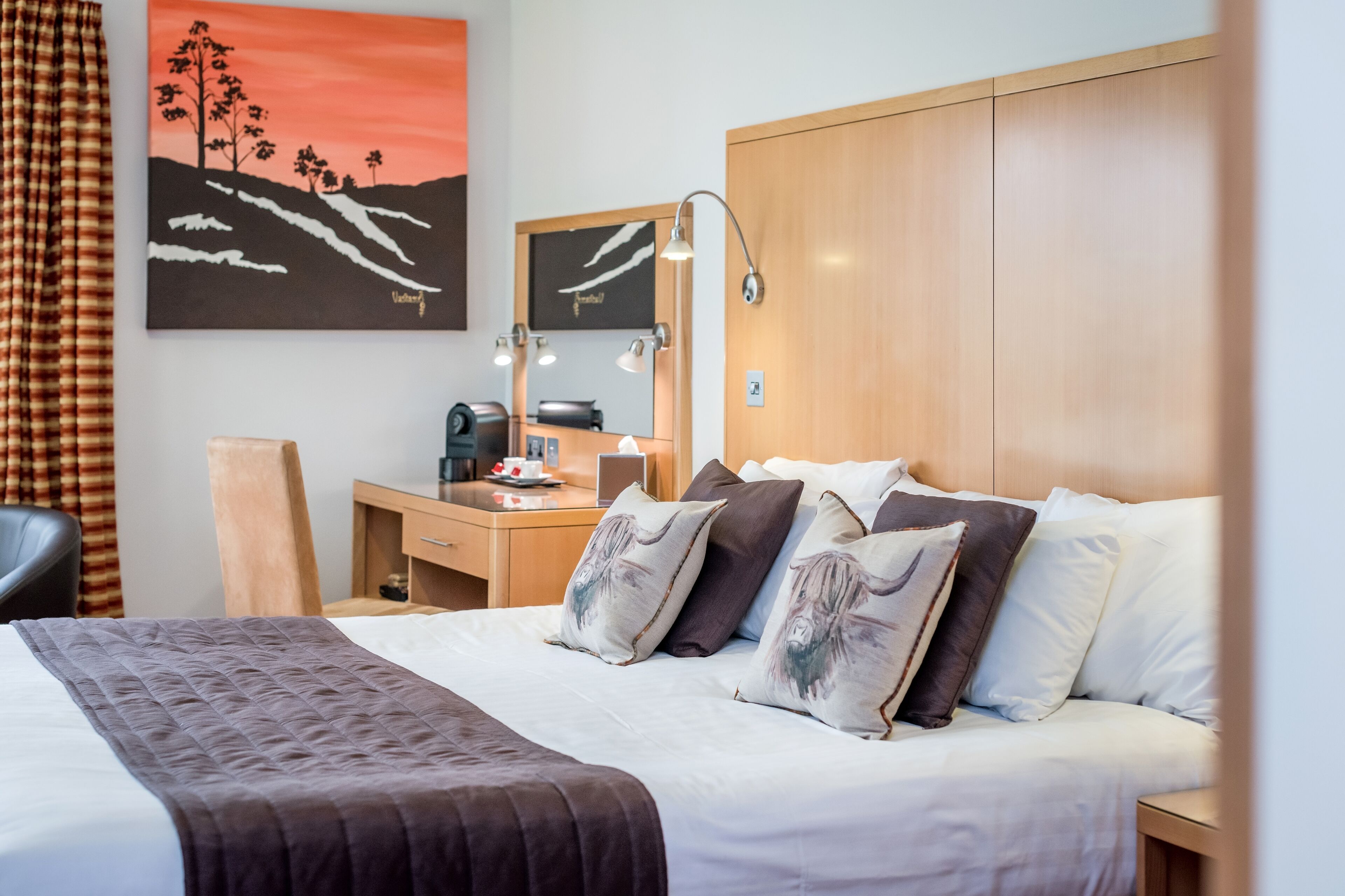 A hotel room with a bed, bedside table, desk, chair, and a painting on the wall.