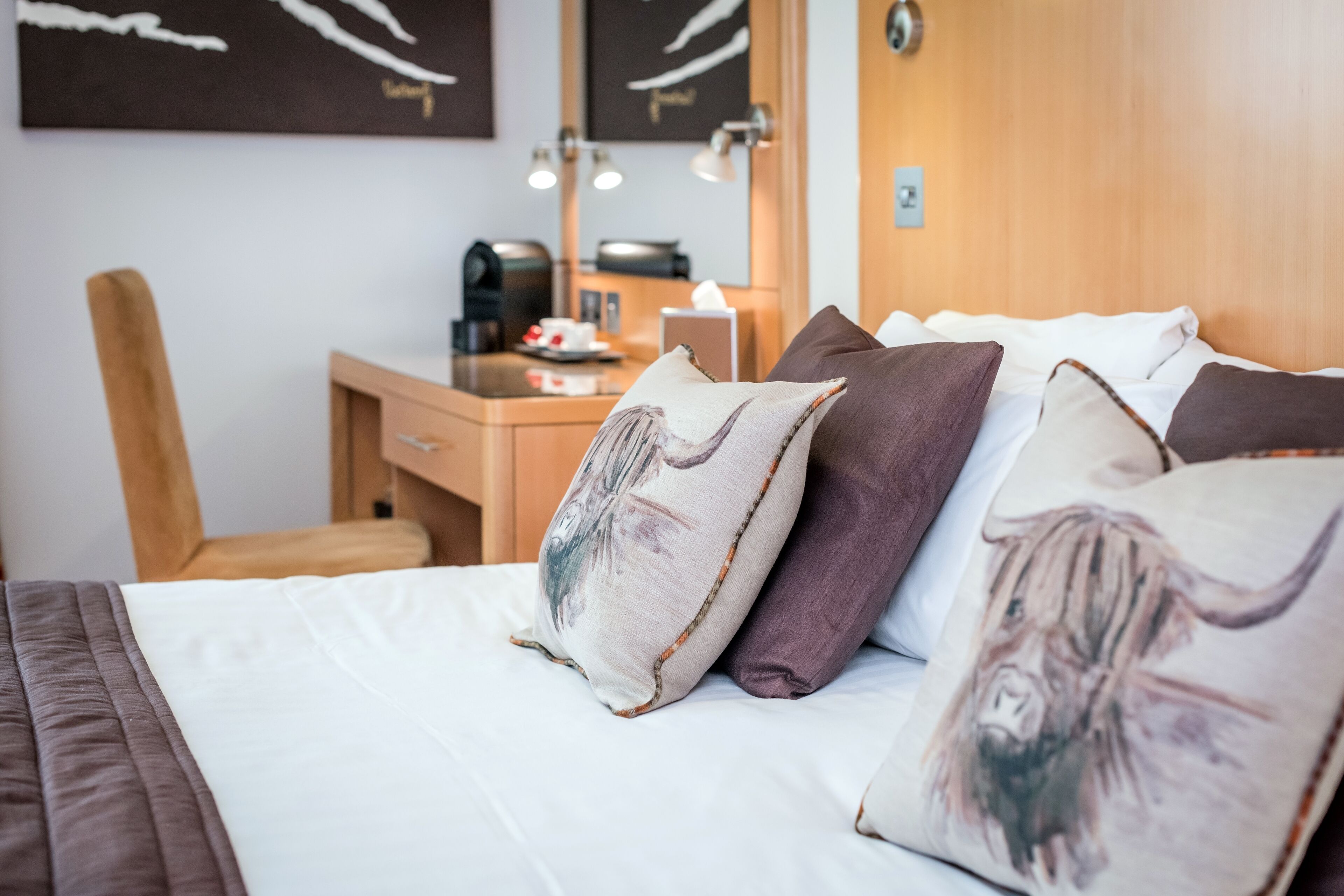 A neatly made bed with multiple pillows, a wooden nightstand with a lamp, and a desk with a coffee maker.