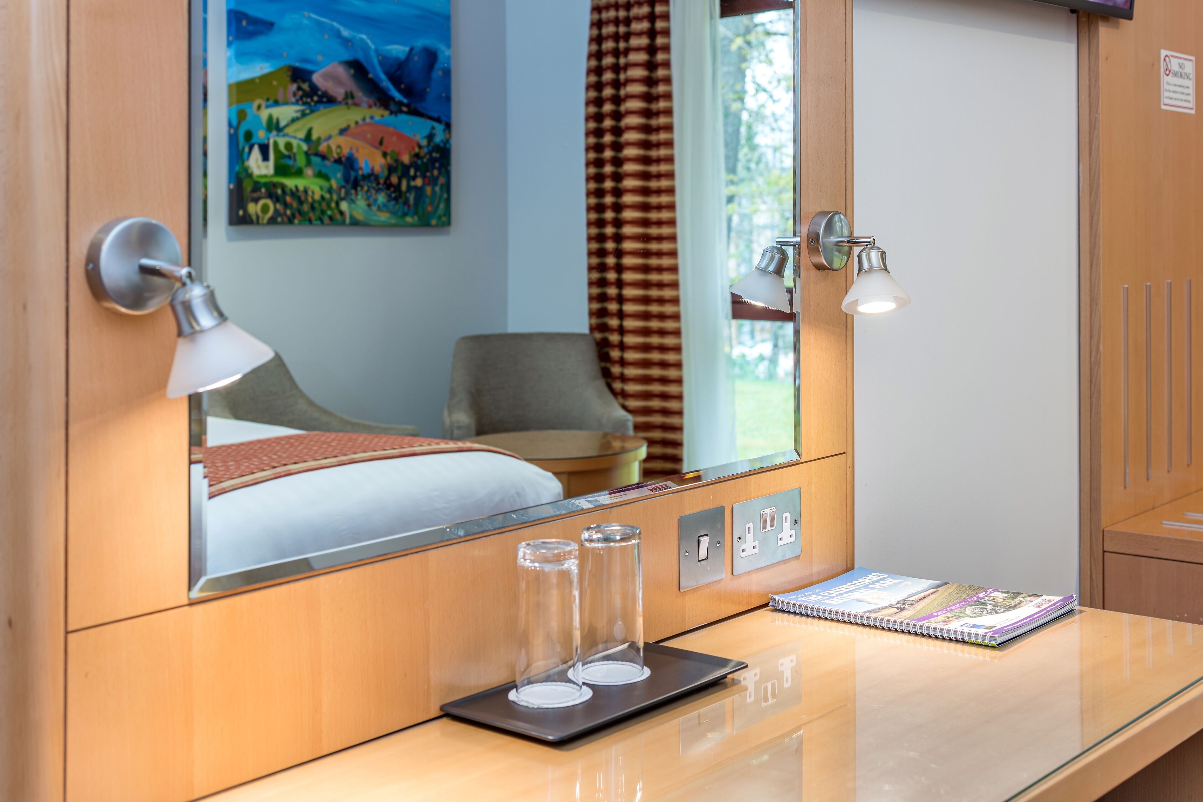 A hotel room with a bed, a bedside table with a lamp, a glass of water, a notebook, and a painting on the wall.