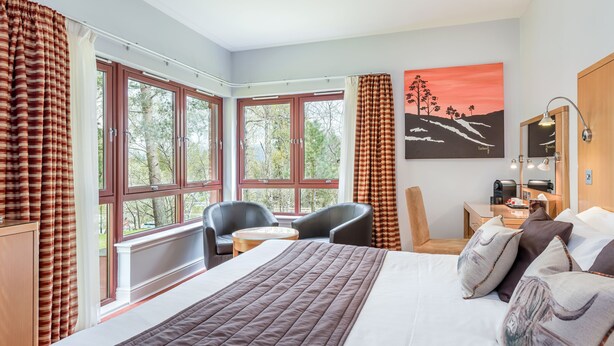 Habitación de hotel con una cama grande, un escritorio con una silla y vistas a árboles a través de amplios ventanales.