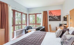 Habitación de hotel con una cama grande, un escritorio con una silla y vistas a árboles a través de amplios ventanales.