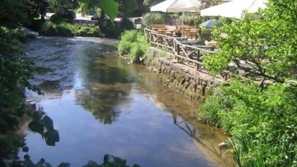 Une rivière traversée par un pont en bois et bordée d’un mur de pierres. Des arbres et de la végétation luxuriante s’y trouvent.