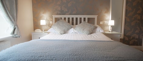 Una cama bien hecha con un edredón azul, dos almohadas y un cabecero blanco. Hay dos mesitas de noche con lámparas y un espejo en una pared.