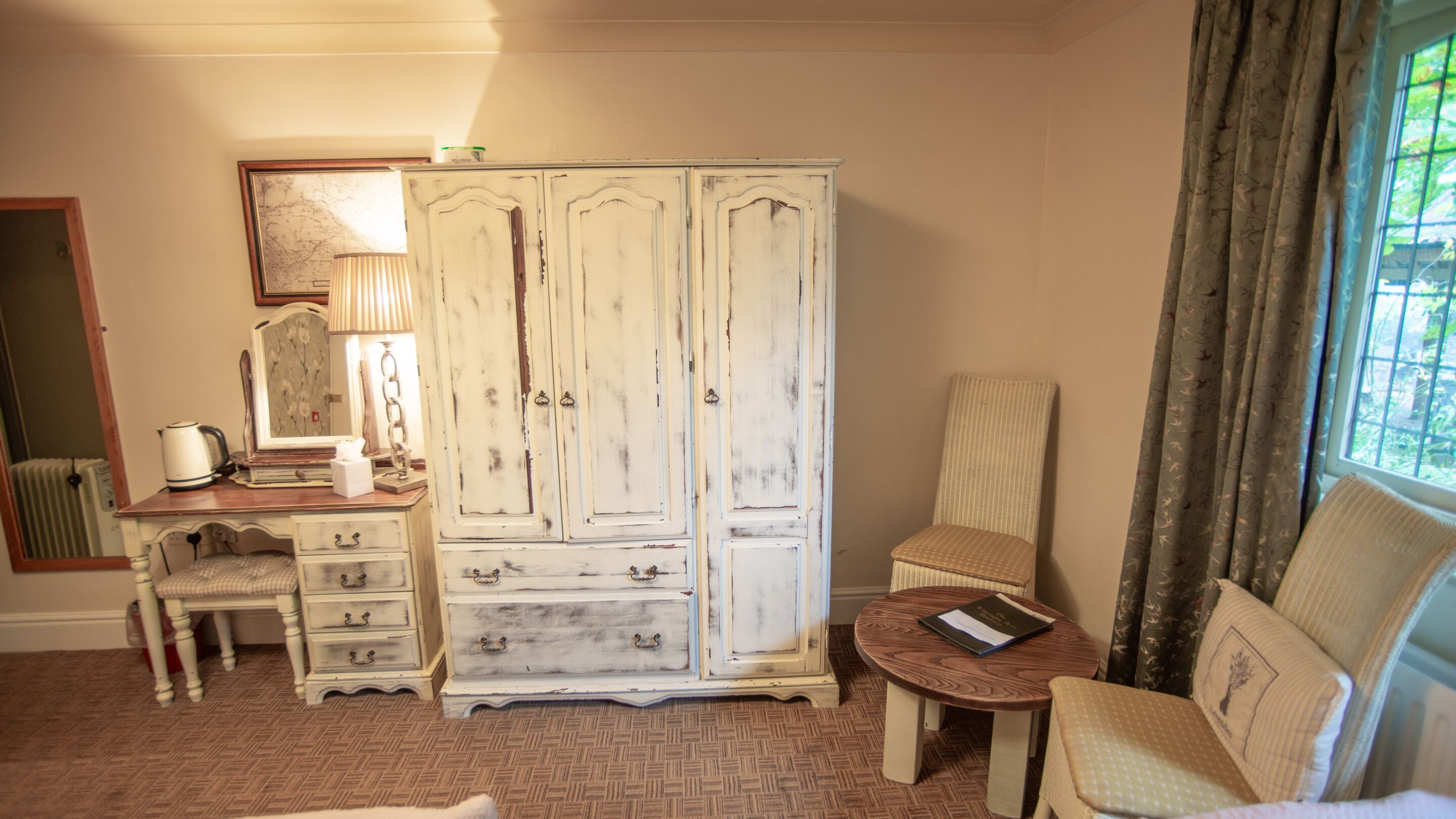 A room with a large white wardrobe, a wooden desk with drawers, a chair, a small round table with a book, and a window with curtains.