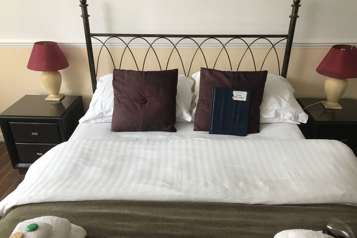 A neatly made bed with white linens, two dark brown pillows, and a small blue book on the nightstand beside it.