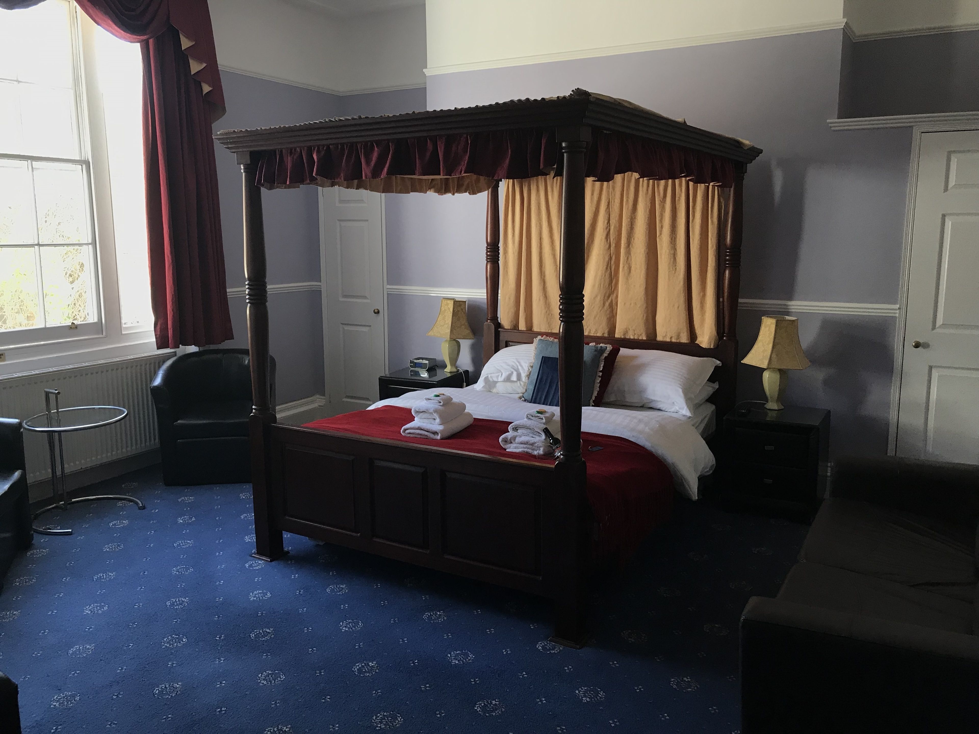 A four-poster bed with a red canopy, a bedside table with a lamp, and a chair in a room with a blue carpet and a window with curtains.