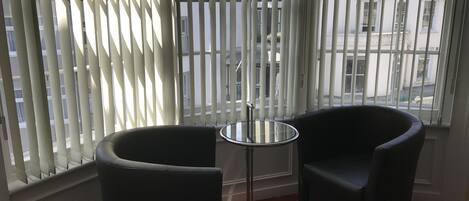 A corner room with two black chairs and a small round table, large windows with blinds, and a red carpet.