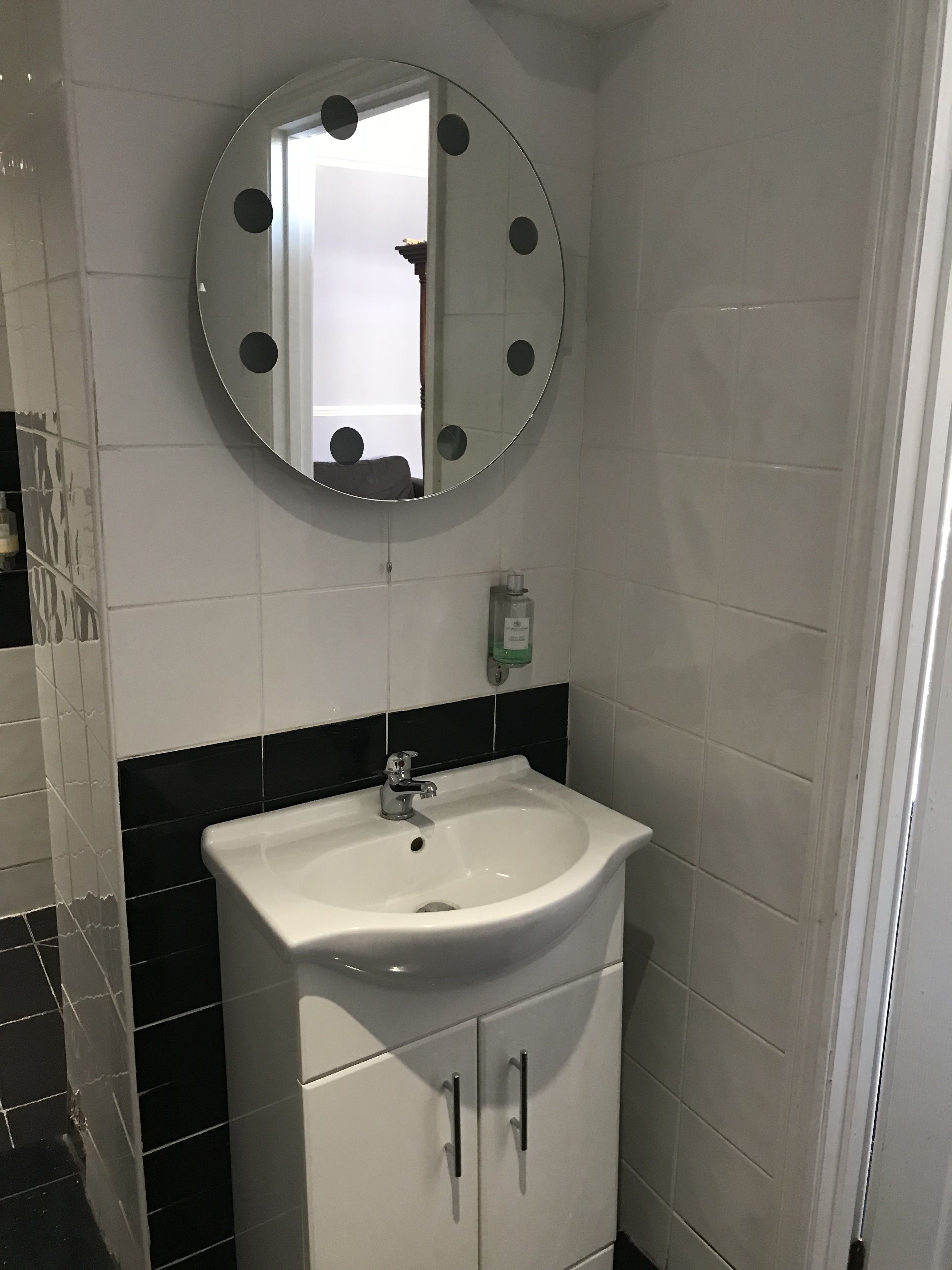Une salle de bain avec un lavabo blanc, des murs carrelés en noir et blanc, un miroir rond orné de pois noirs et un distributeur de savon vert.