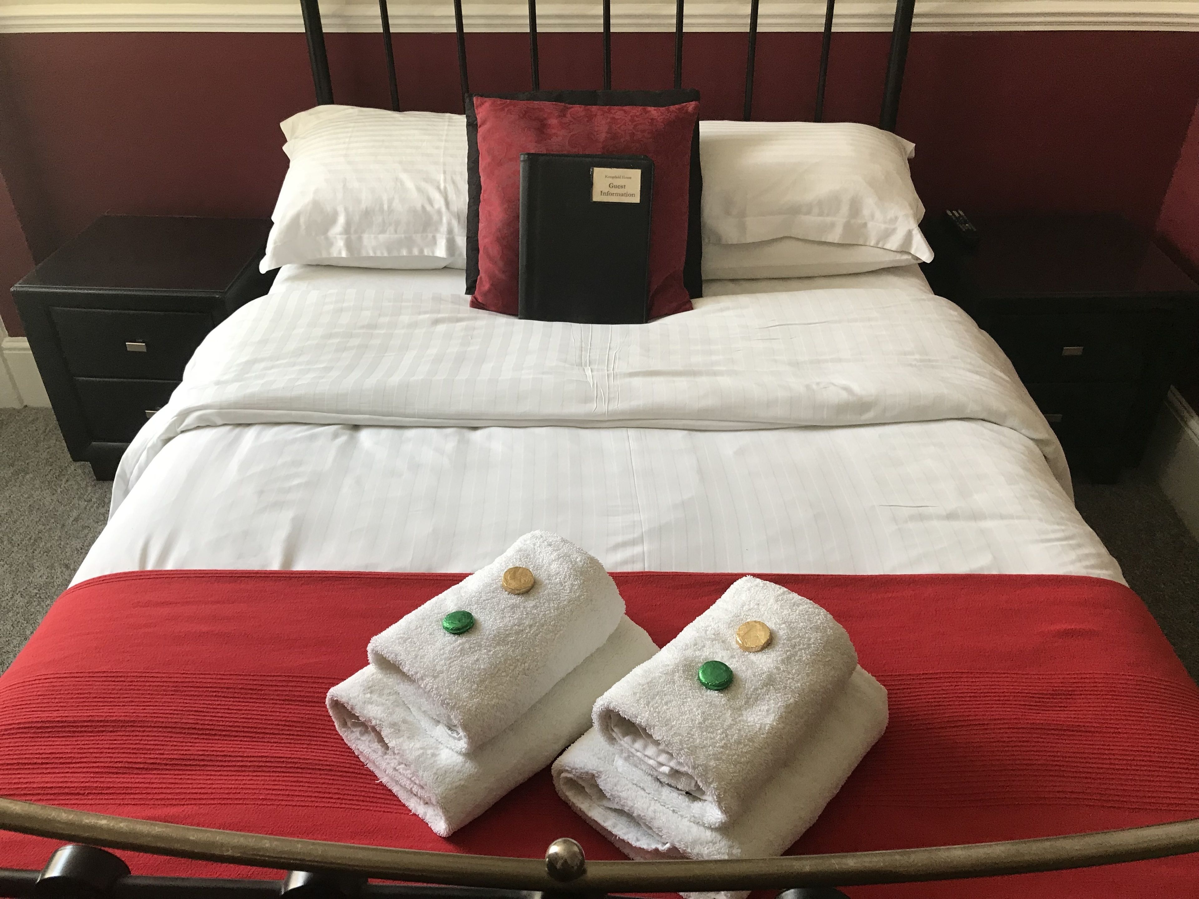 A neatly made bed with white linens, a red bedspread, and two folded towels with green and yellow buttons.