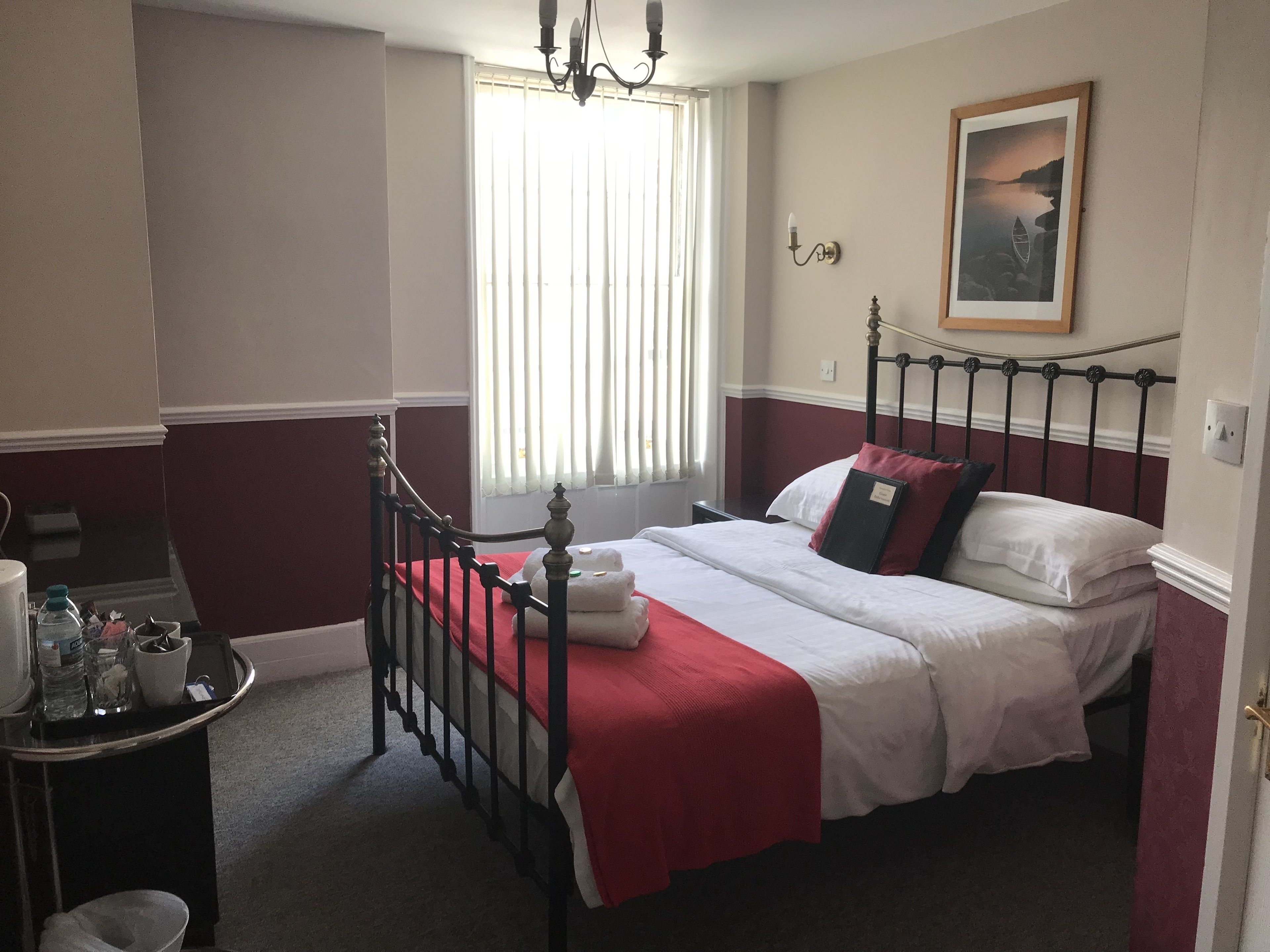 Un dormitorio con cama, mesita de noche, ventana con persianas, candelabro y un cuadro en la pared.