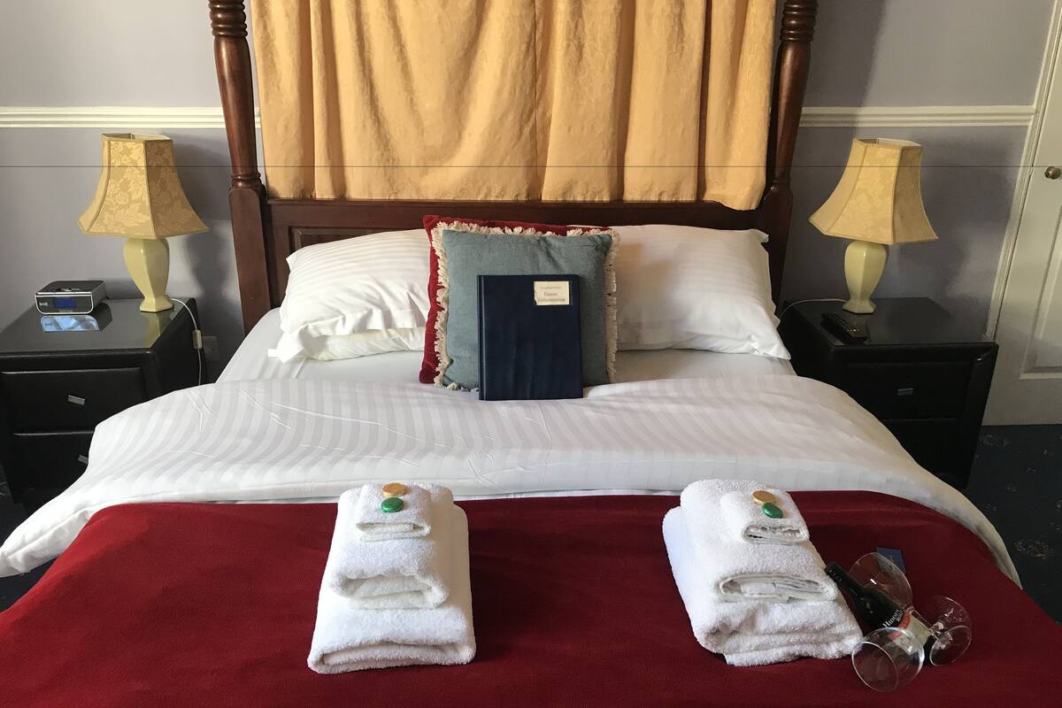A neatly made bed with a red bedspread, two folded towels with bath salts, a bottle of alcohol, and a glass on the side.