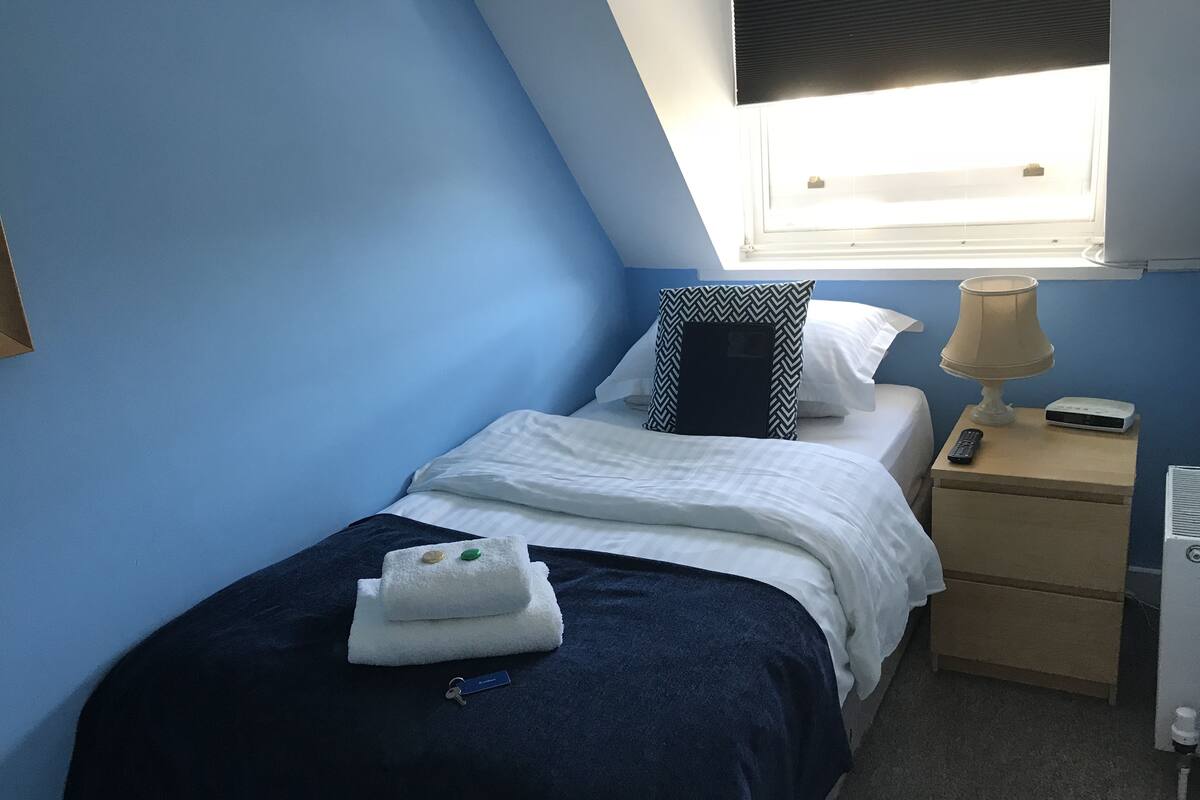 A bedroom with a bed, a nightstand, a lamp, a remote control, and a window with blinds.