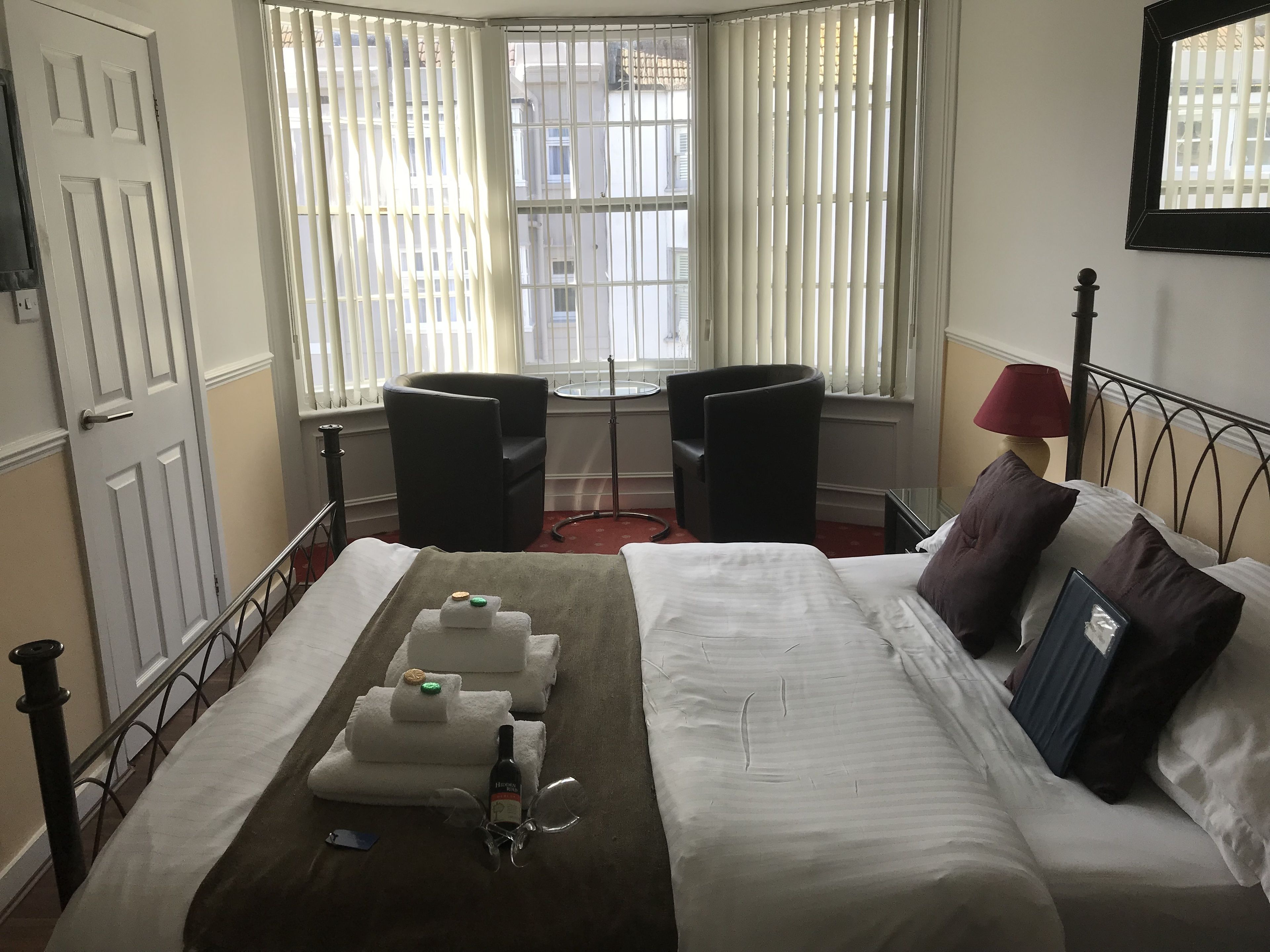 A hotel room with a bed, two chairs, a small table, and a window with blinds.