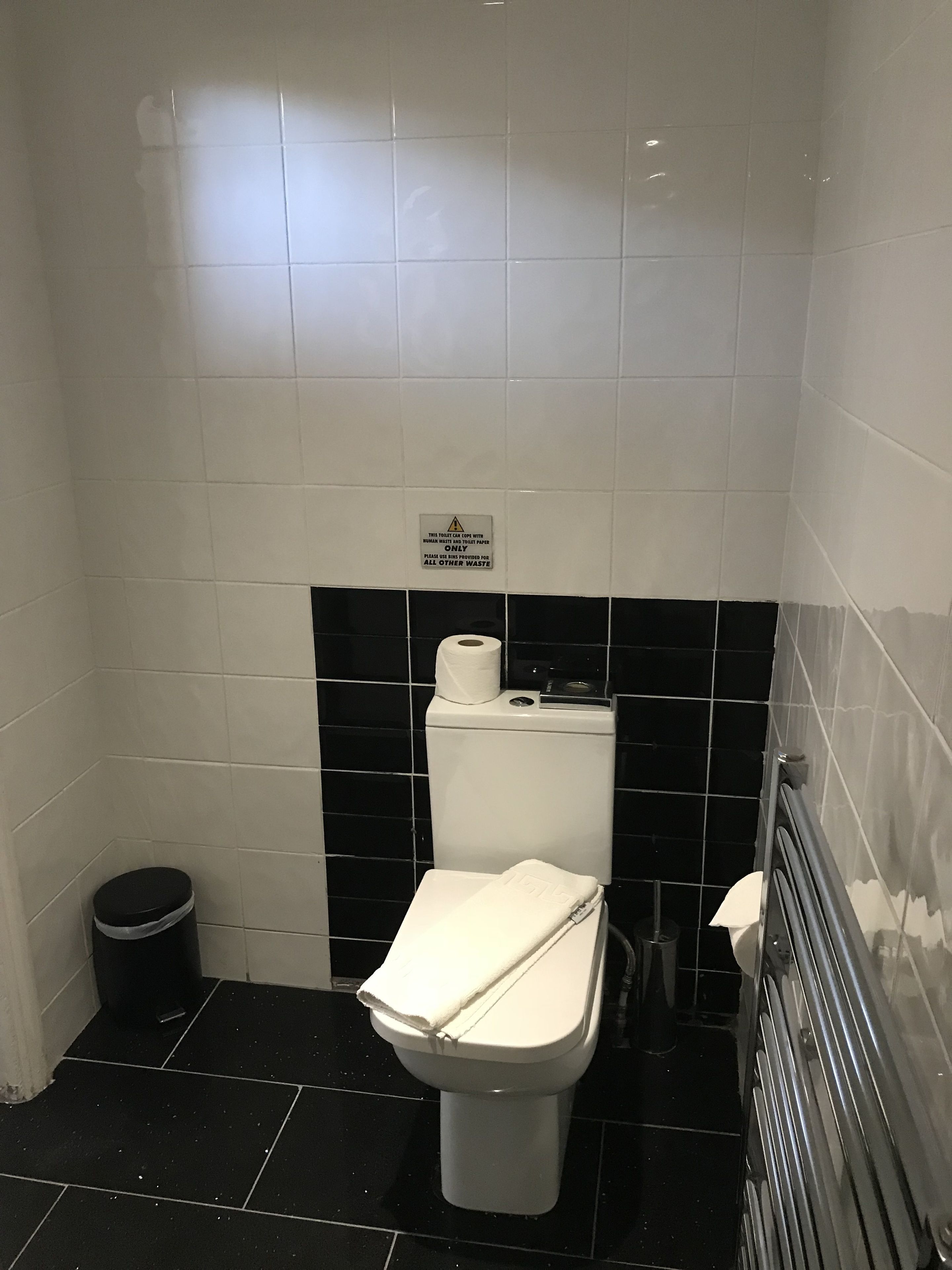 Un baño pequeño con azulejos, un inodoro, un cesto de basura y un letrero que indica tirar el papel higiénico por el inodoro.