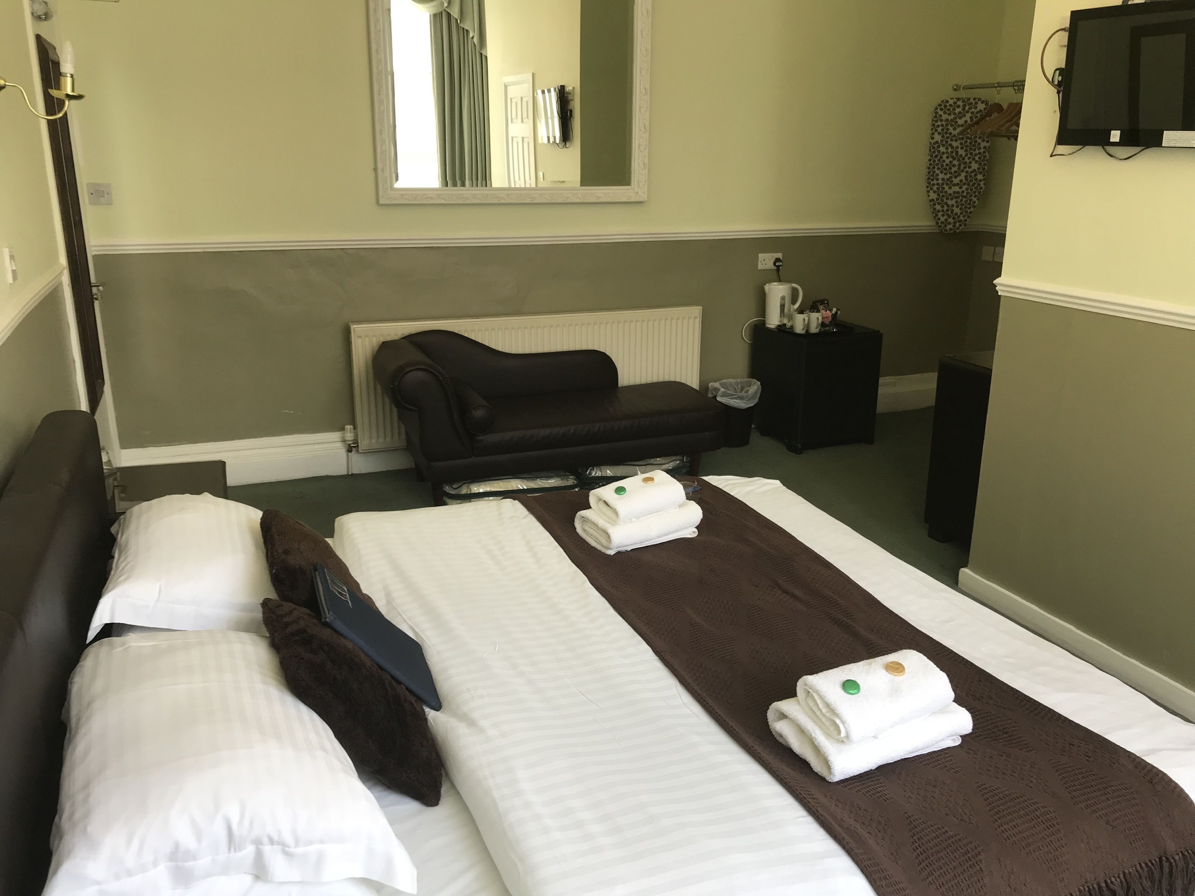 A hotel room with two beds, a black sofa, a mirror, a radiator, a small table with toiletries, and a television on the wall.