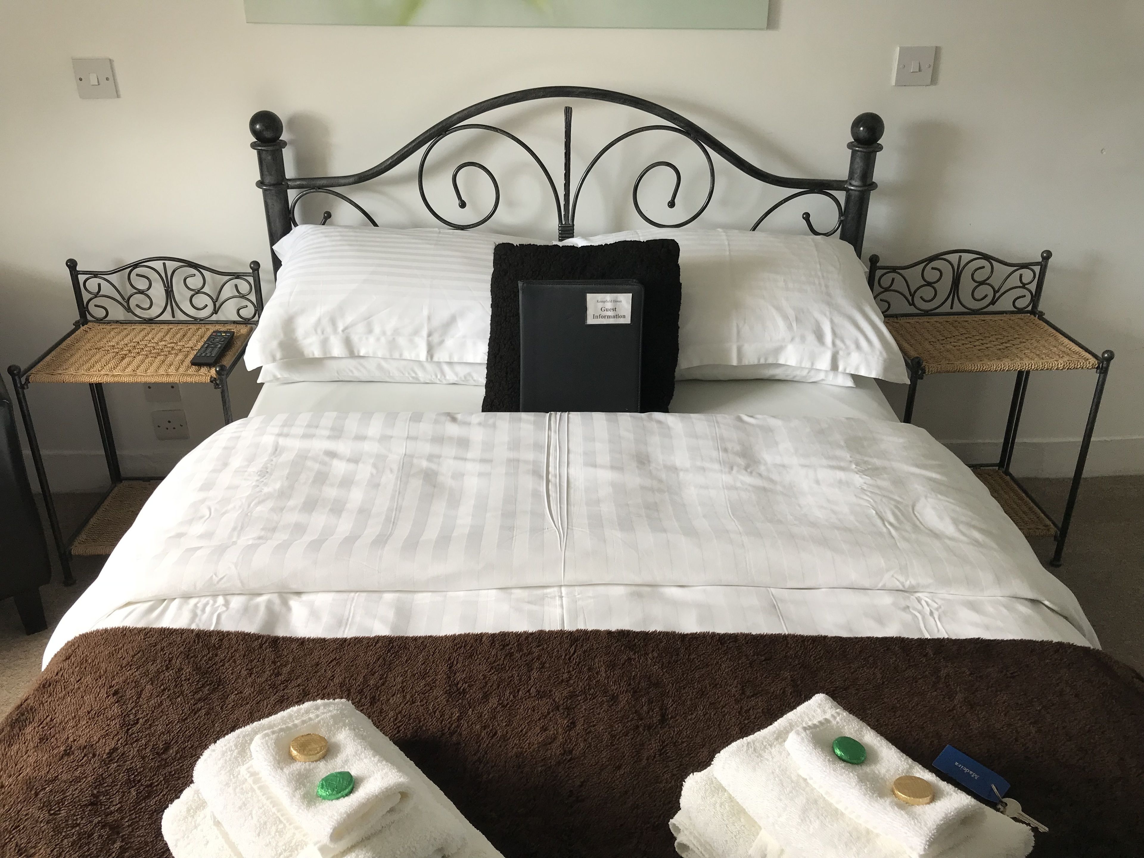 A neatly made bed with white linens, a brown throw blanket, and two towels with bath salts on a bedside table.