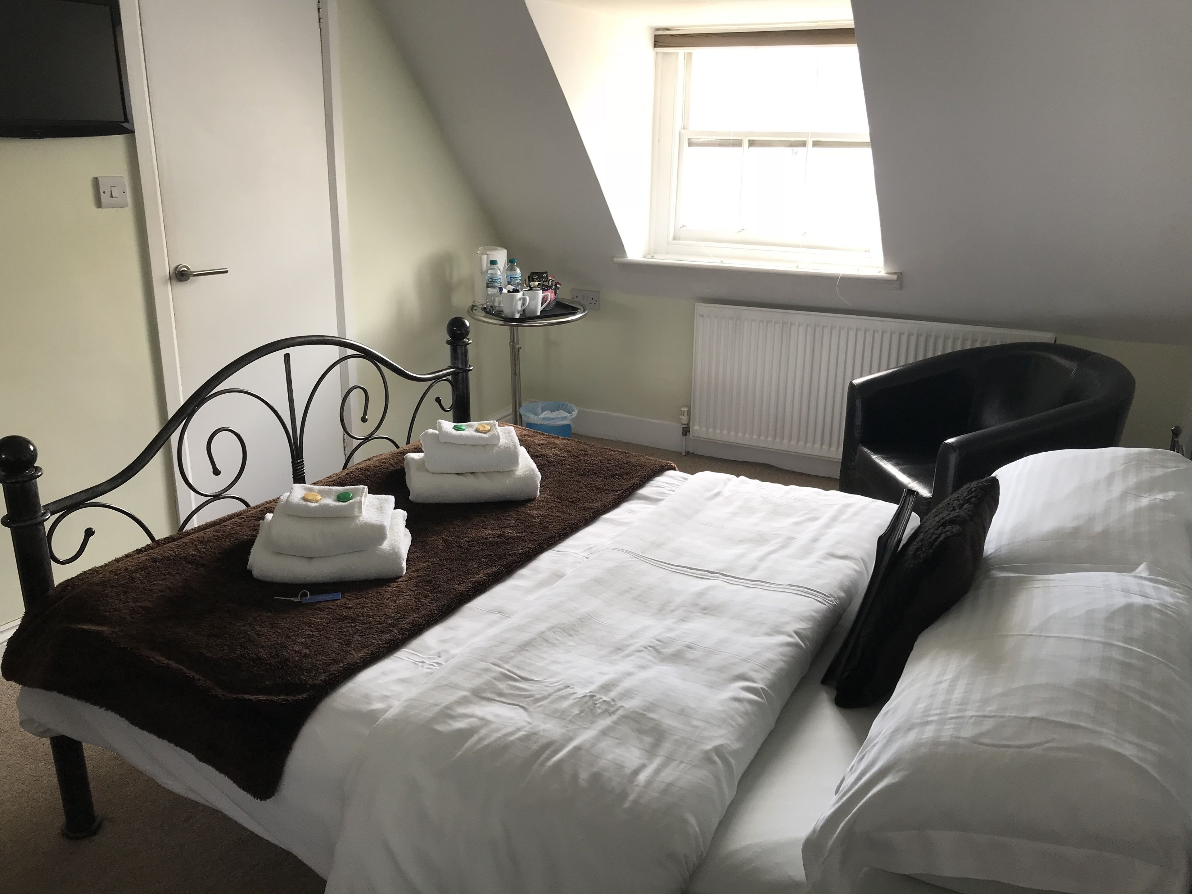 A neatly made bed with white linens and a brown throw blanket, a black metal headboard, a small bedside table with a lamp, and a window with blinds.
