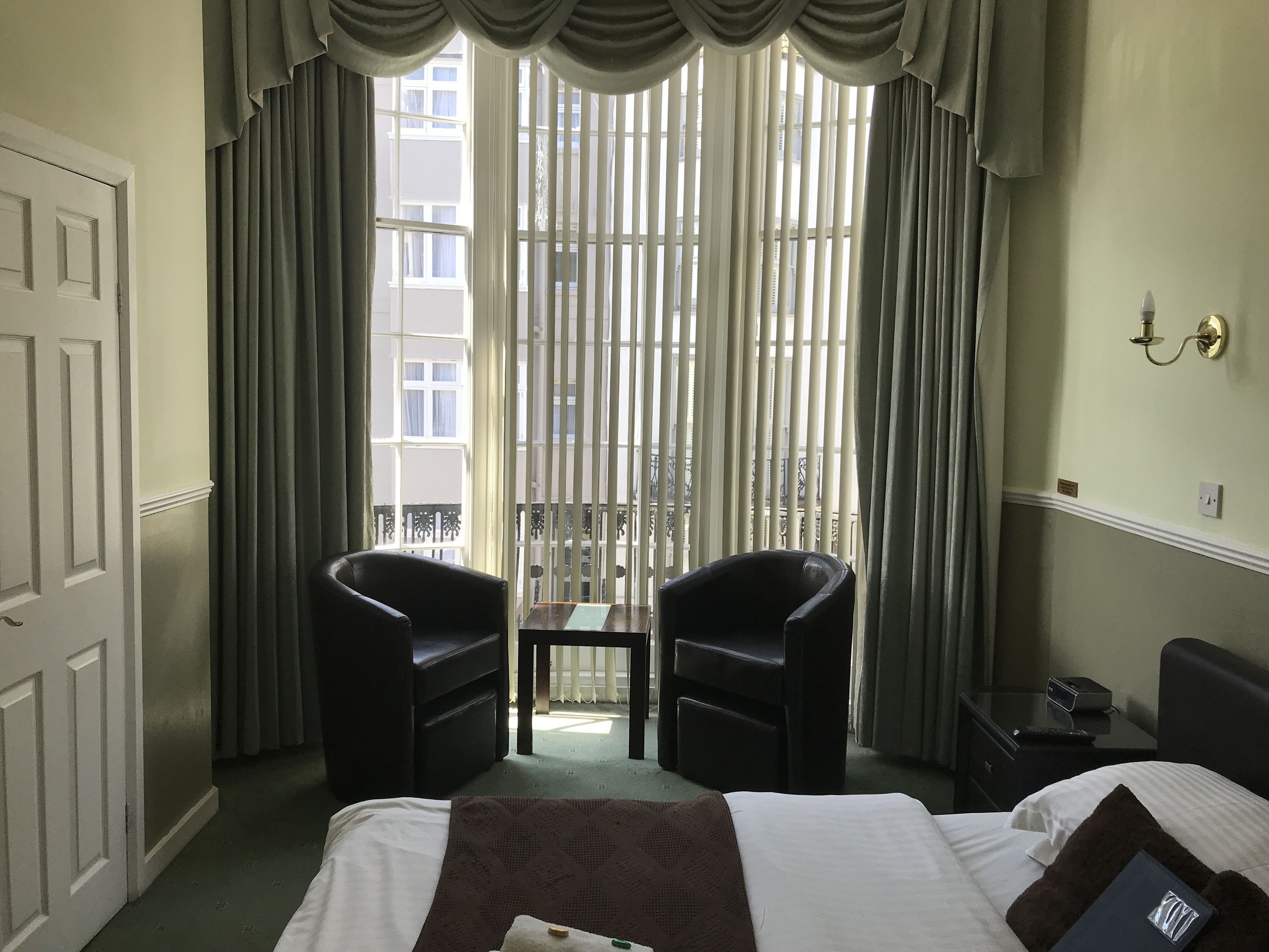 A hotel room with a bed, two armchairs, a small table, and a view through floor-to-ceiling windows.