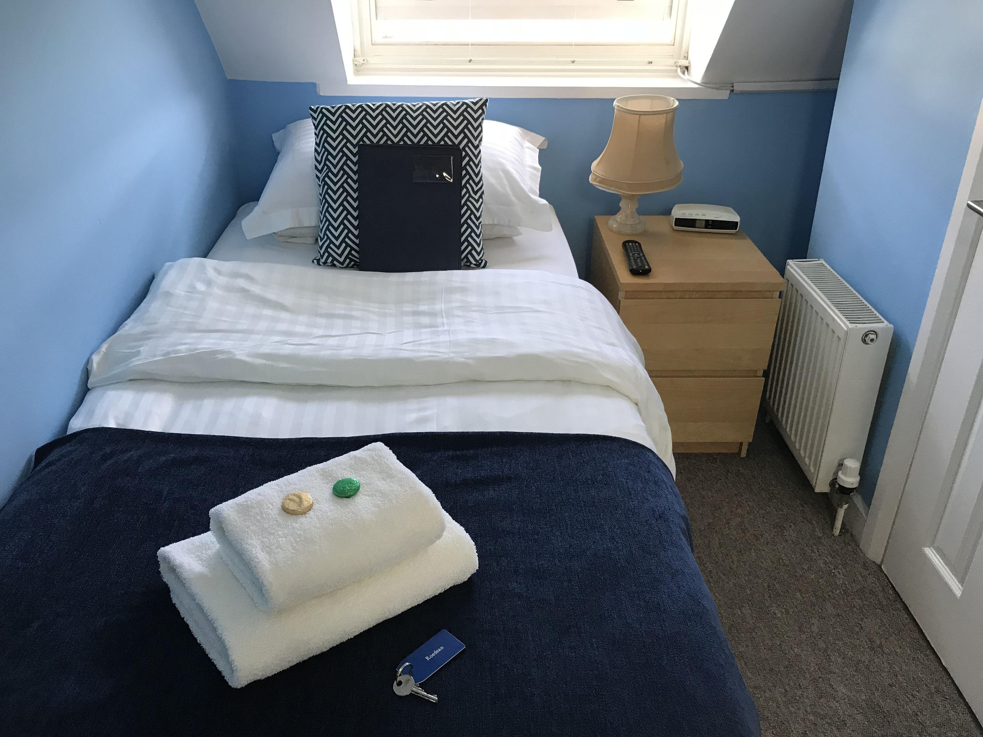 A neatly made bed with white linens, a blue blanket, and a folded towel with a keychain. There is a black and white patterned pillow, a bedside lamp, and a small wooden nightstand with a remote control.