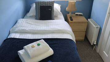 Una cama bien hecha con sábanas blancas, un edredón azul y una toalla doblada con un llavero. Hay un cojín de estampado blanco y negro, una lámpara de noche y una mesita de noche de madera con un control remoto.
