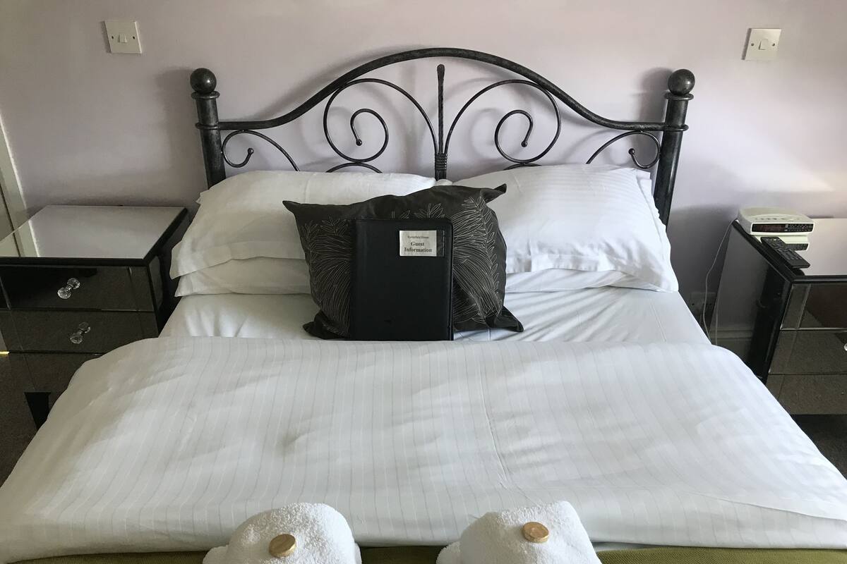 A neatly made bed with white linens, a black pillow, and a black book on the side. There are two nightstands with a lamp and a phone on one, and a small table with a remote control on the other.
