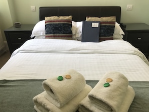 A neatly made bed with white linens, two decorative pillows, a blue information booklet, and two folded towels with green and gold bath salts.