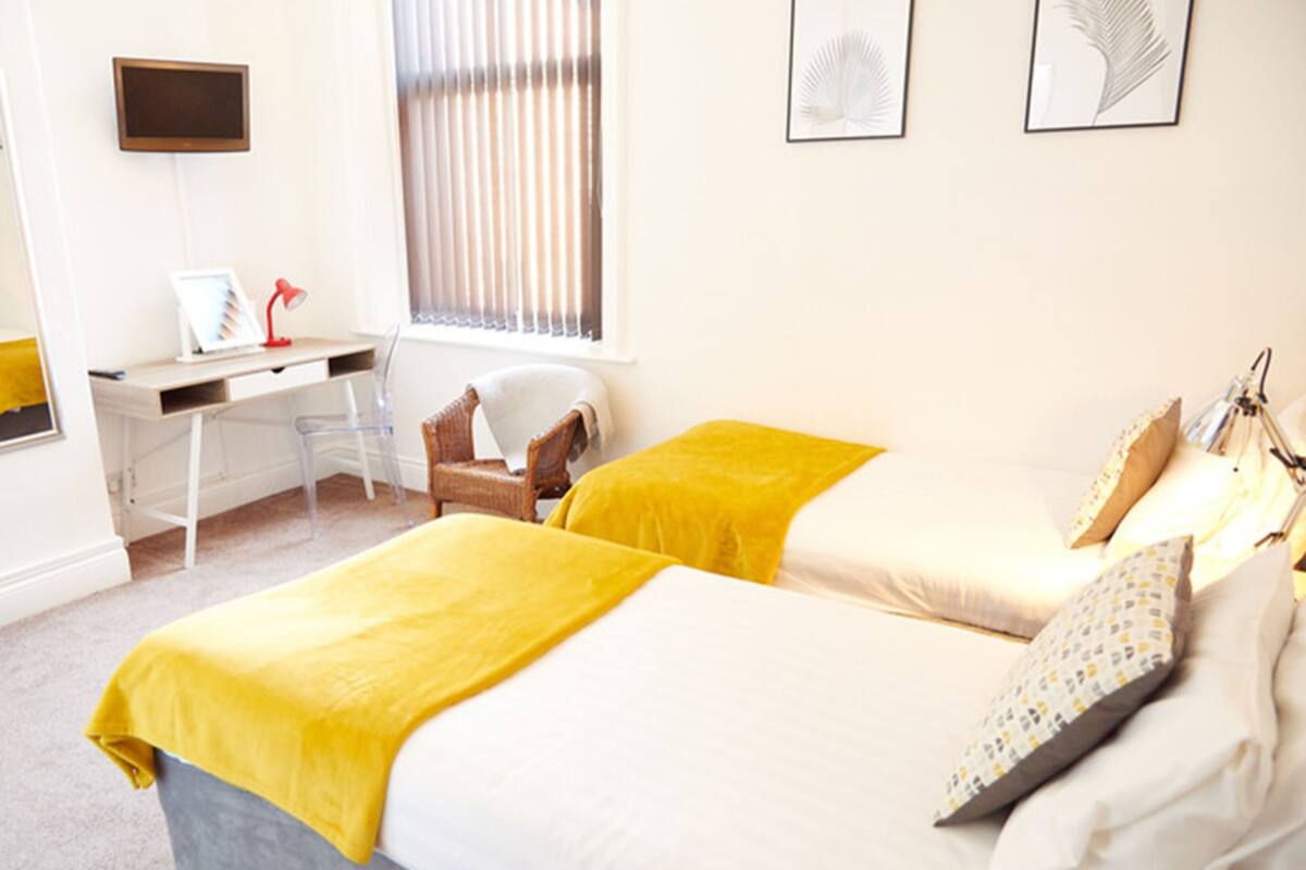 A bedroom with two beds, a desk, a chair, a TV, and a window with blinds.
