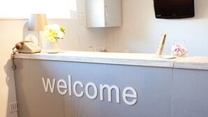 Un mostrador de recepción con un cartel de 'welcome', un teléfono, un florero con flores y un televisor montado en la pared.