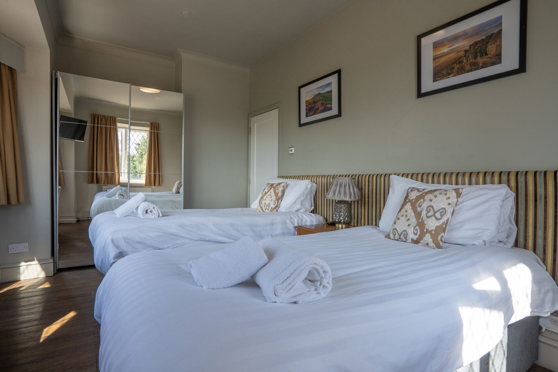 A hotel room with two beds, a mirror, a window, and two framed pictures on the wall.