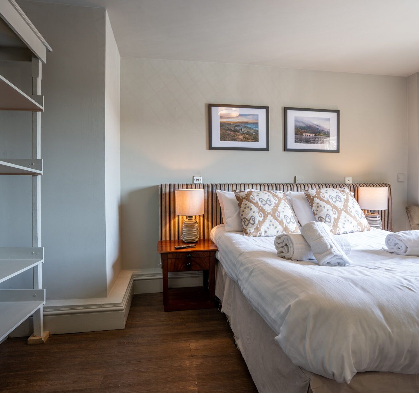 Un dormitorio con una cama, dos lámparas de noche, una mesita de noche, piso de madera y dos cuadros en la pared.