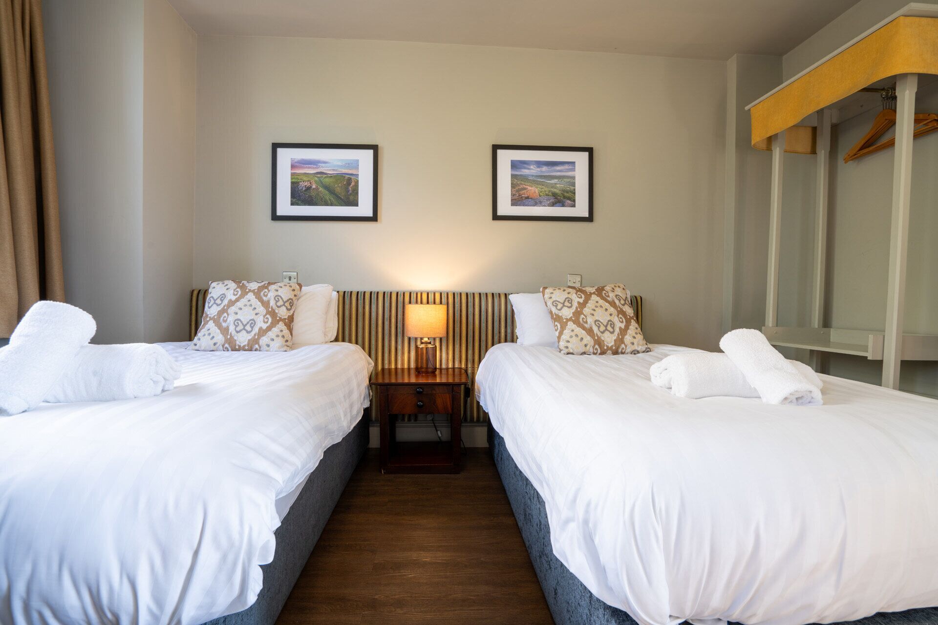 Habitación de hotel con dos camas, una mesita de noche, una lámpara y dos cuadros en la pared.