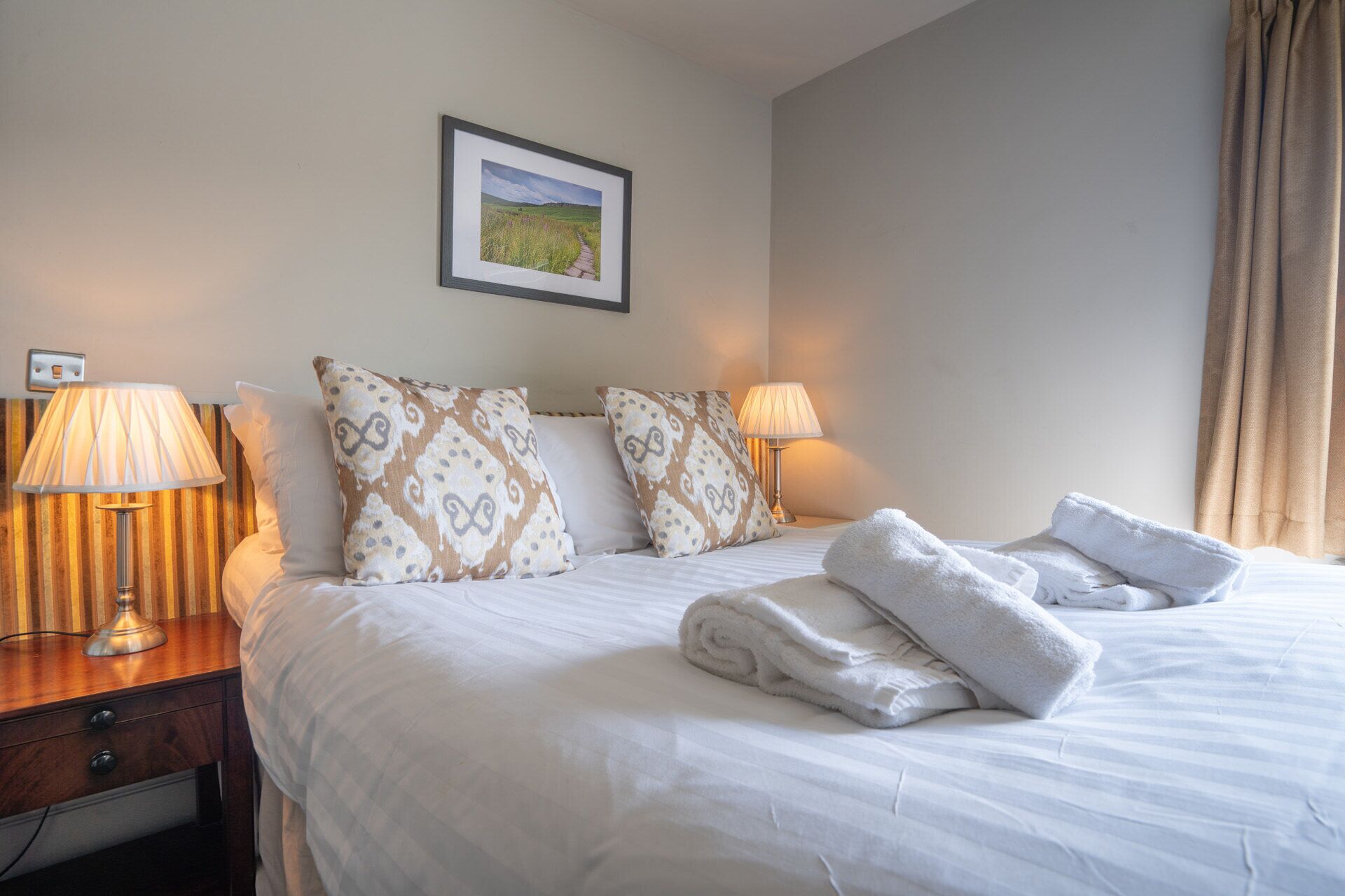 A neatly made bed with white linens, two bedside lamps, patterned pillows, a framed picture on the wall, and a window with curtains.