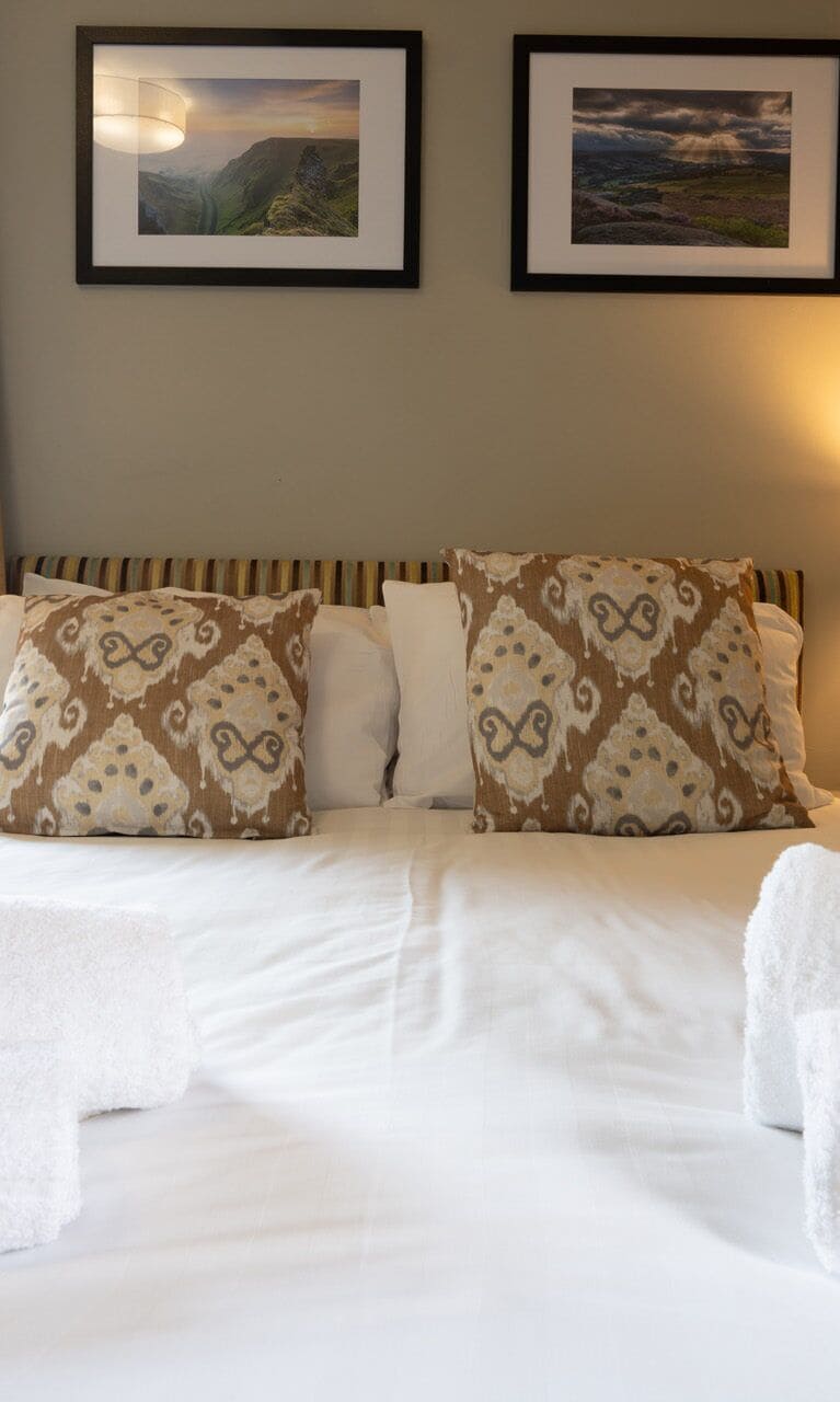 Uma cama com almofadas estampadas, um cobertor branco e duas fotos de paisagens emolduradas na parede.