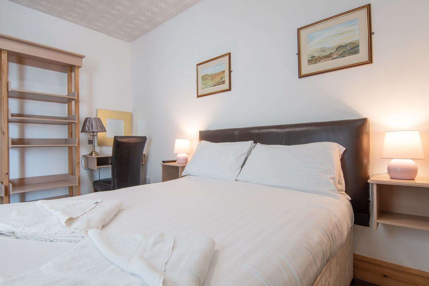 A neatly made bed with white linens, two bedside lamps, a wooden chair, a desk with a lamp, and two framed pictures on the wall.