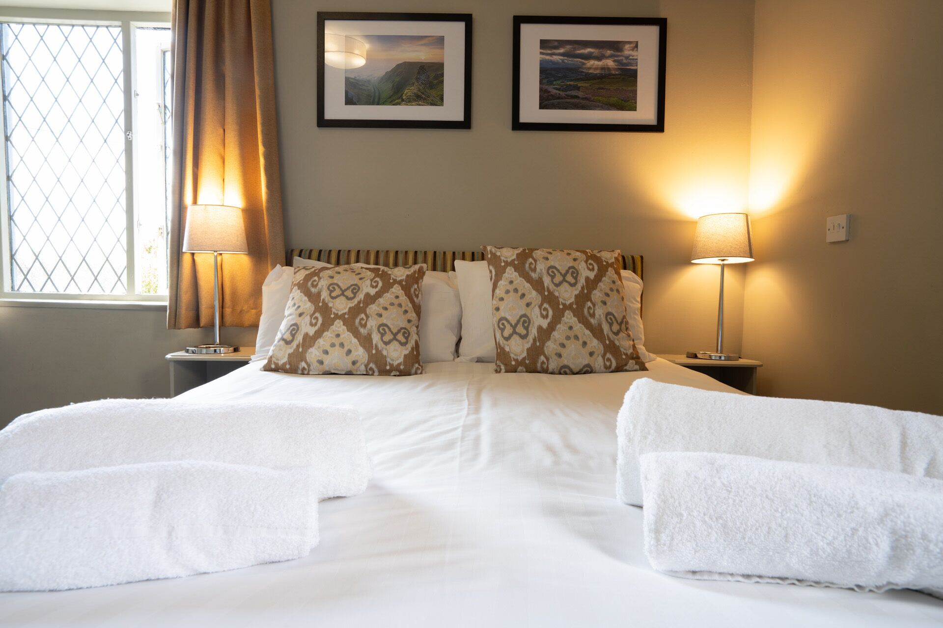A hotel room with a bed, two framed pictures on the wall, a window with curtains, and two bedside lamps.