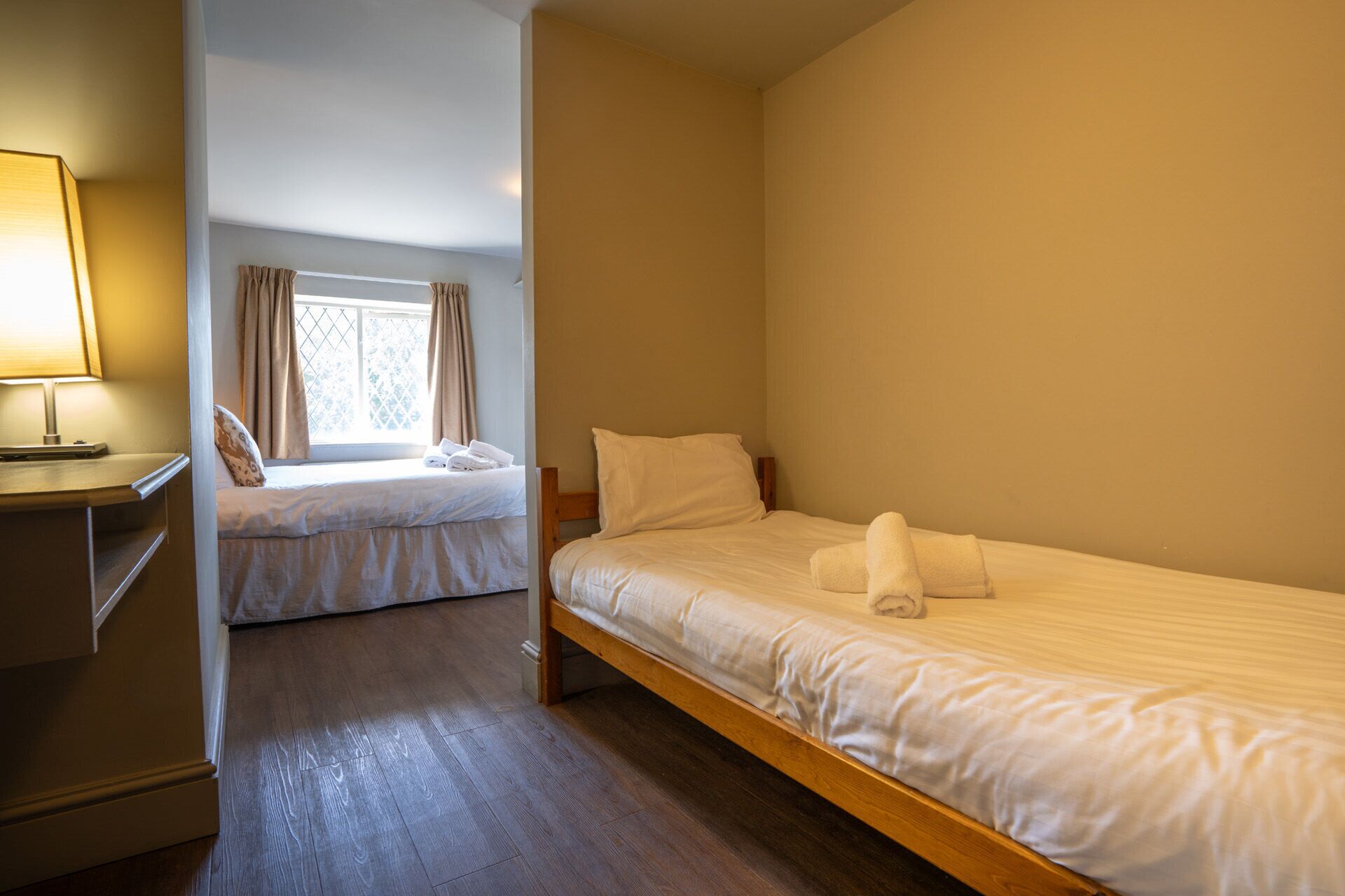 Un dormitorio con dos camas, una ventana con cortinas, una mesita de noche con lámpara y suelo de madera.