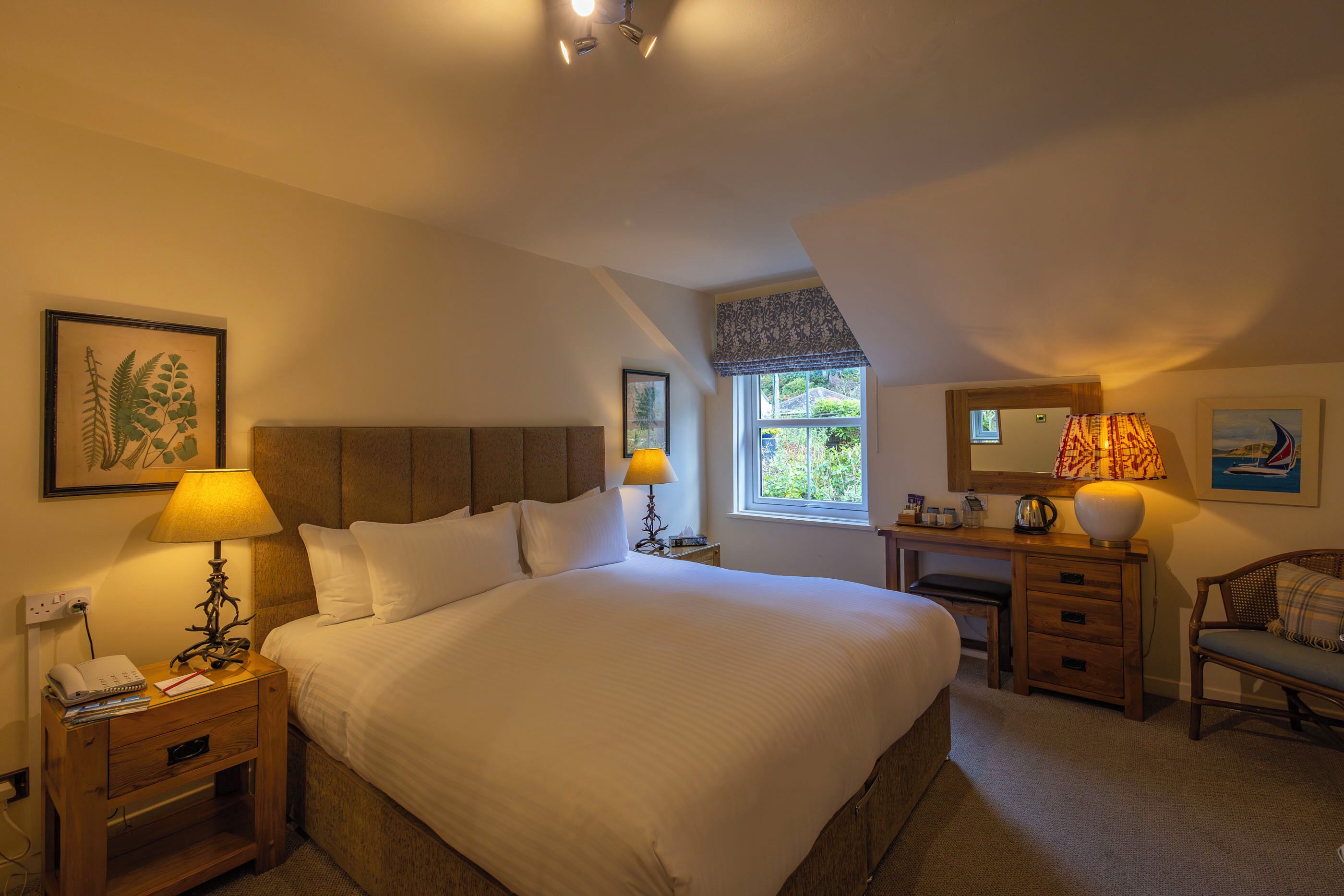 A hotel room with a large bed, two bedside lamps, a wooden nightstand, a desk with a chair, a window with a view of greenery, and a painting on the wall.