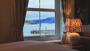 Ein Zimmer mit großem Fenster und Blick auf See und Berge. Ein Bett mit gestreifter Bettdecke und eine Lampe auf einem Nachttisch.