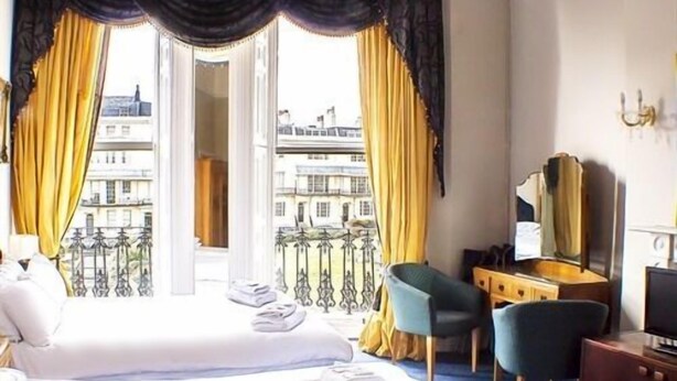 A hotel room with two beds, a desk, a chair, a TV, and a balcony with yellow curtains.