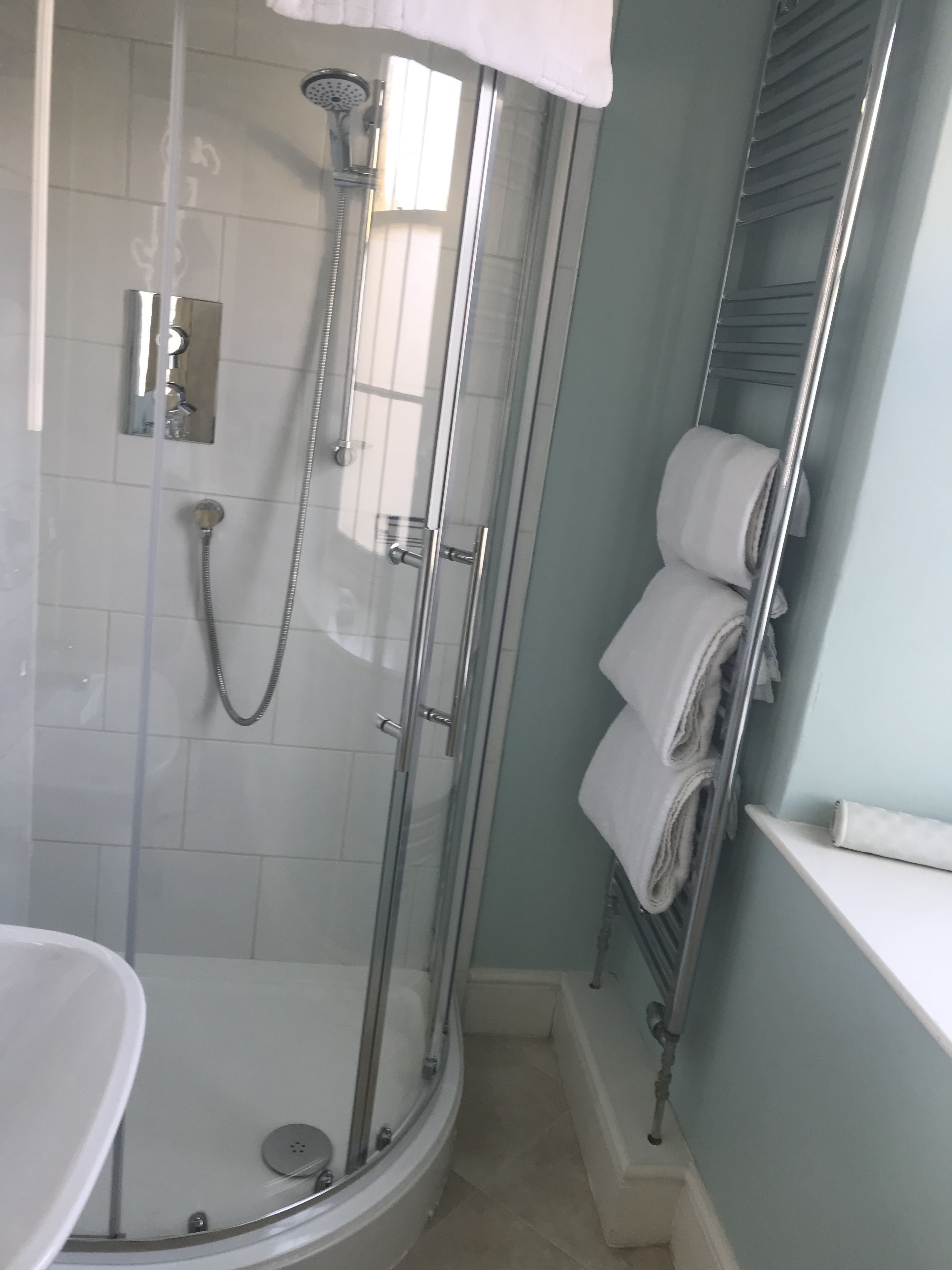 A glass-enclosed shower with a handheld showerhead, a wall-mounted faucet, and a separate handheld showerhead. There is a white bathtub with a glass door, and a white towel rack with folded towels.