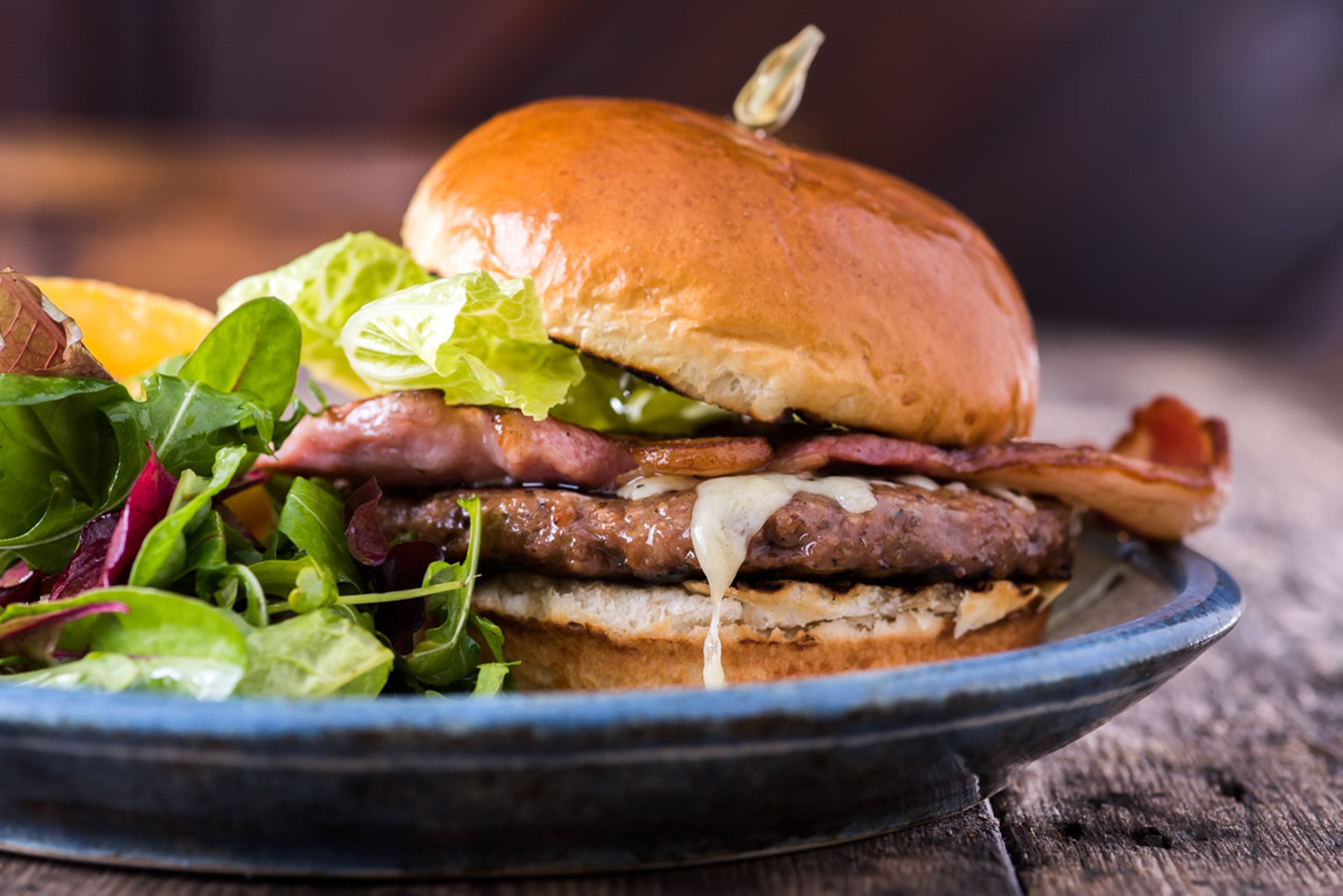 A burger with lettuce, bacon, and melted cheese on a bun, served on a plate with a side of greens.