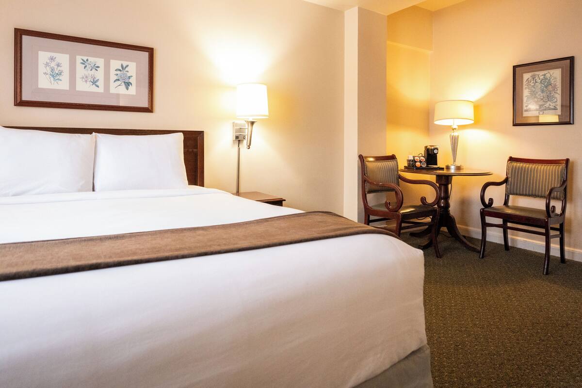A hotel room with a large bed, two bedside lamps, a small table with chairs, and framed artwork on the walls.