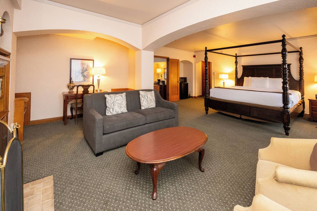 A hotel room with a four-poster bed, a sofa, a coffee table, and a desk with a lamp.