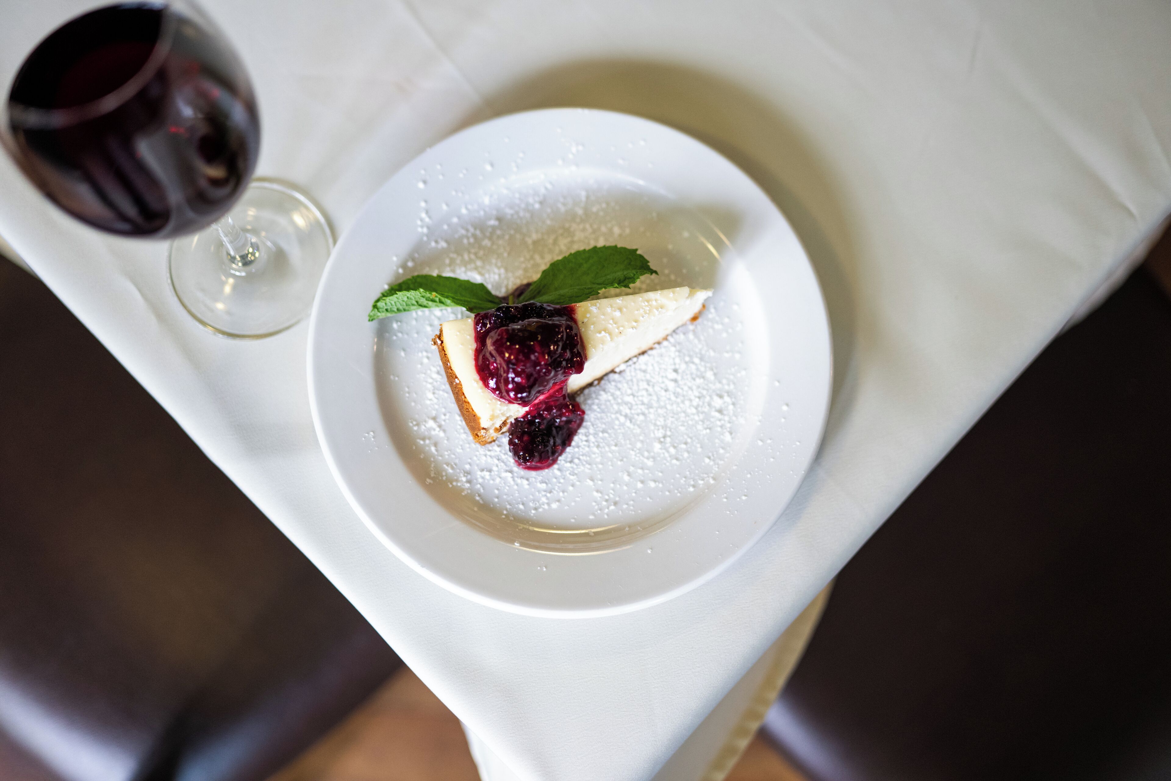 Un plato de postre con una porción de pastel de queso, compota de frutos rojos y hojas de menta, acompañado por una copa de vino tinto sobre un mantel blanco.