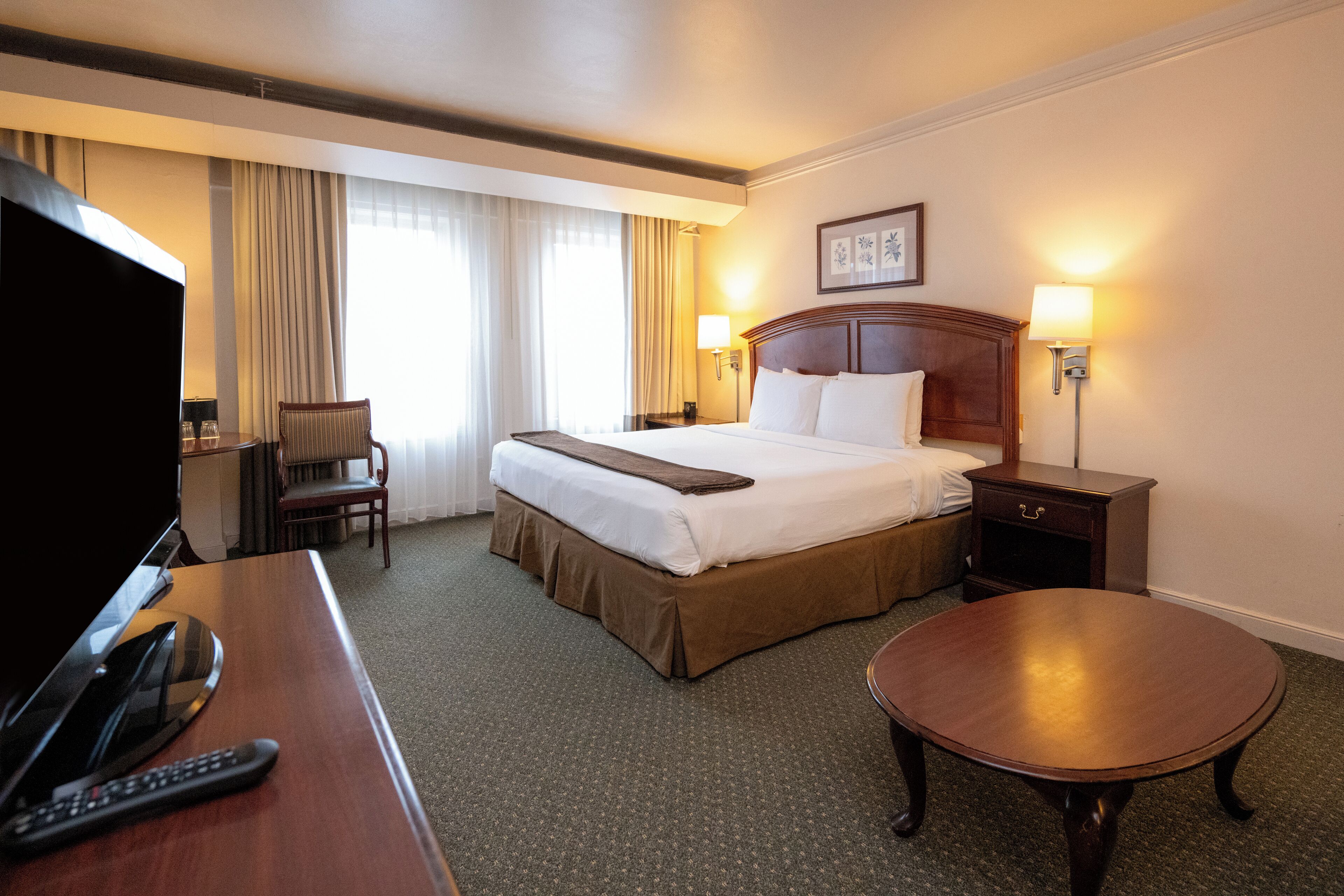Habitación de hotel con una cama grande, un escritorio con televisor y control remoto, una silla, una mesita de noche con lámparas y una mesa redonda.