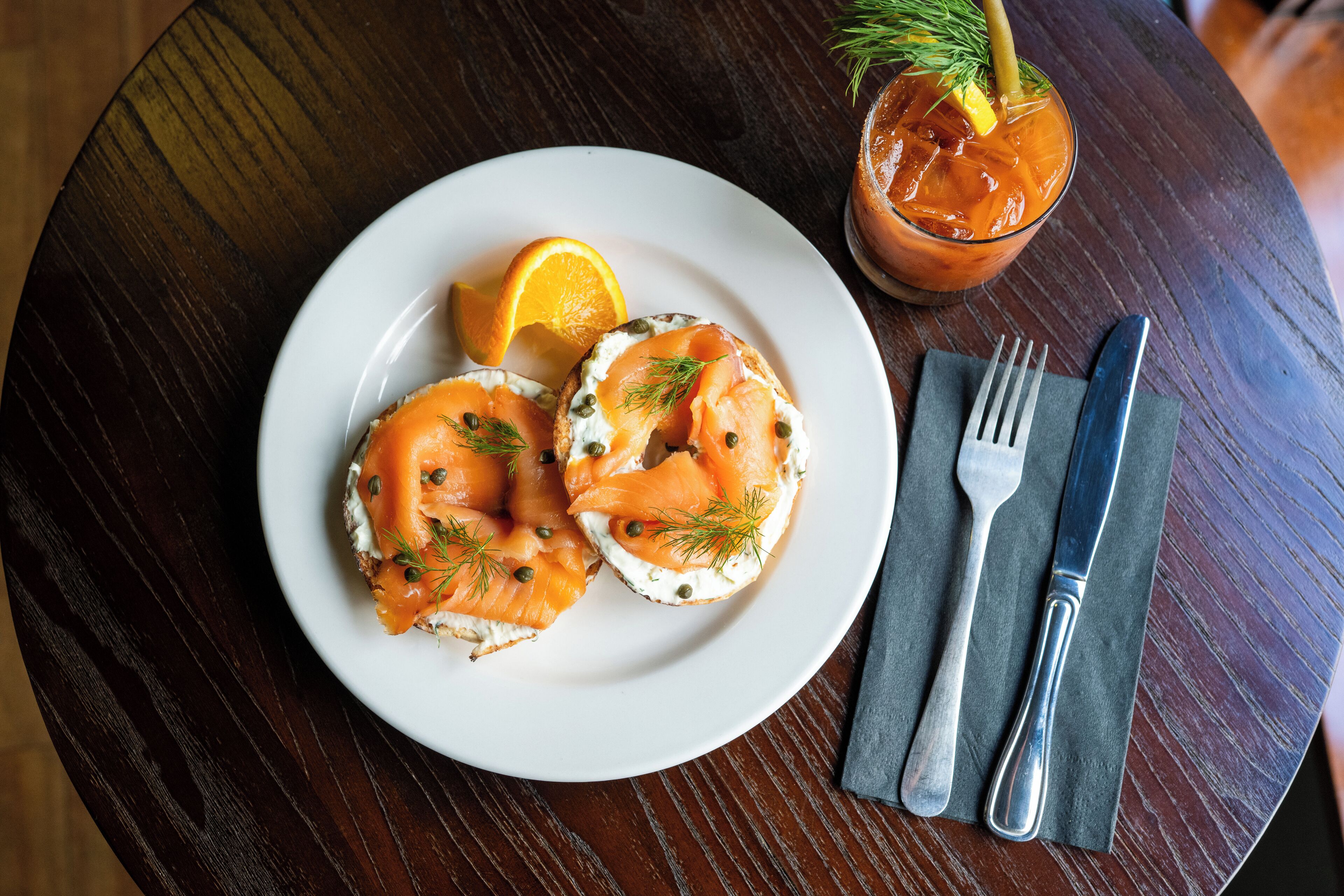 Une assiette avec deux tartines garnies de saumon fumé et de câpres, une tranche d’orange et un verre de thé glacé avec une rondelle de citron et une branche d’aneth.